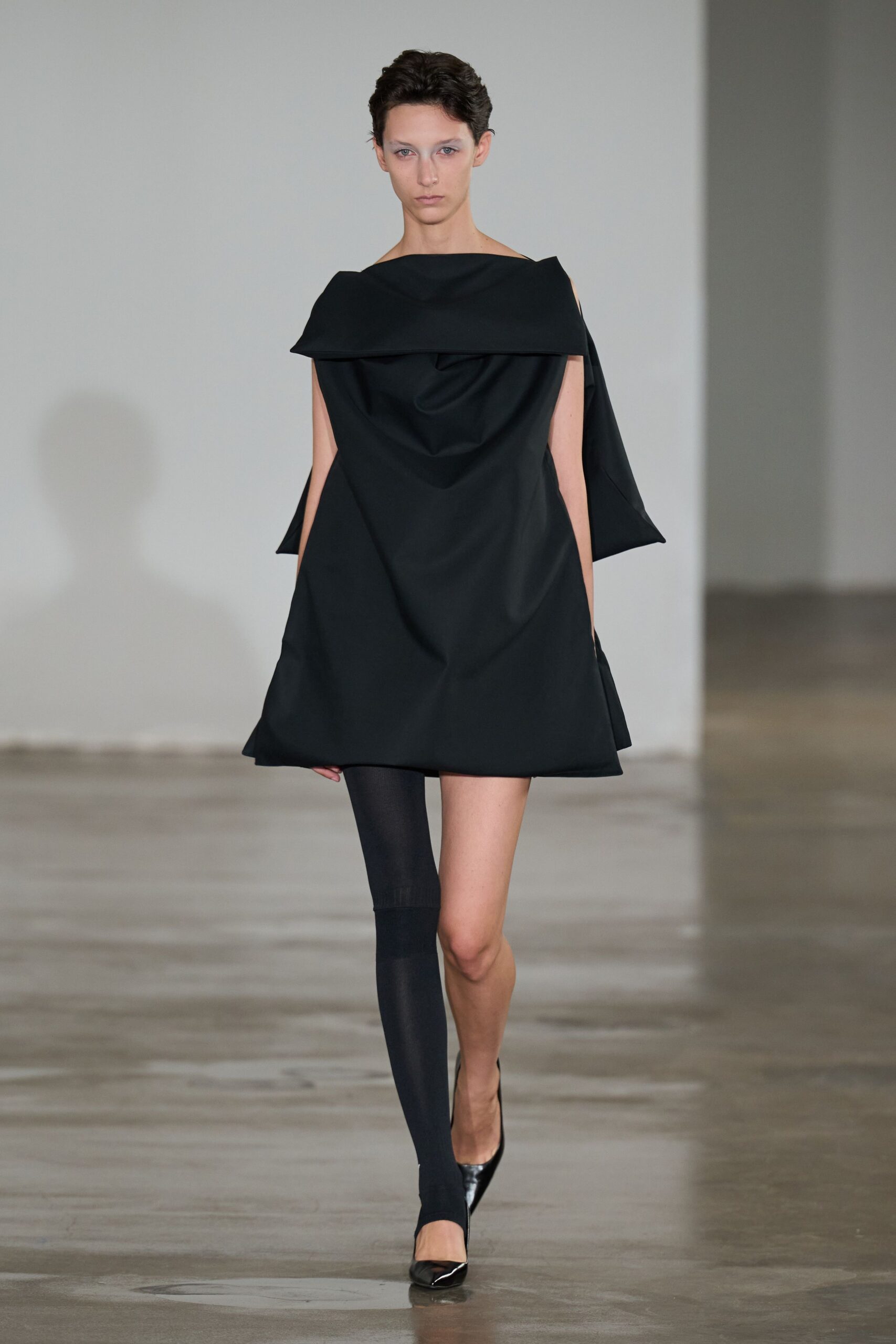 Black structured mini dress with a wide folded collar and cocoon drape. Single opaque black leg, ending in a stirrup under the foot. Black pointed pumps with a sharp patent gleam at the toe. For crossing a museum atrium when the floor still smells of fresh sealant.