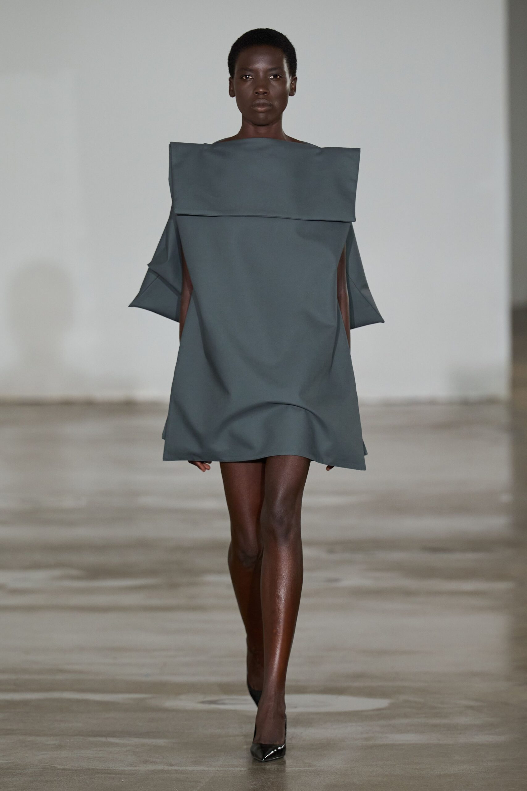 Slate neoprene shift dress with a wide folded yoke and open sides. Black patent pumps, sharply pointed. Crossing an empty museum atrium before the doors unlock.