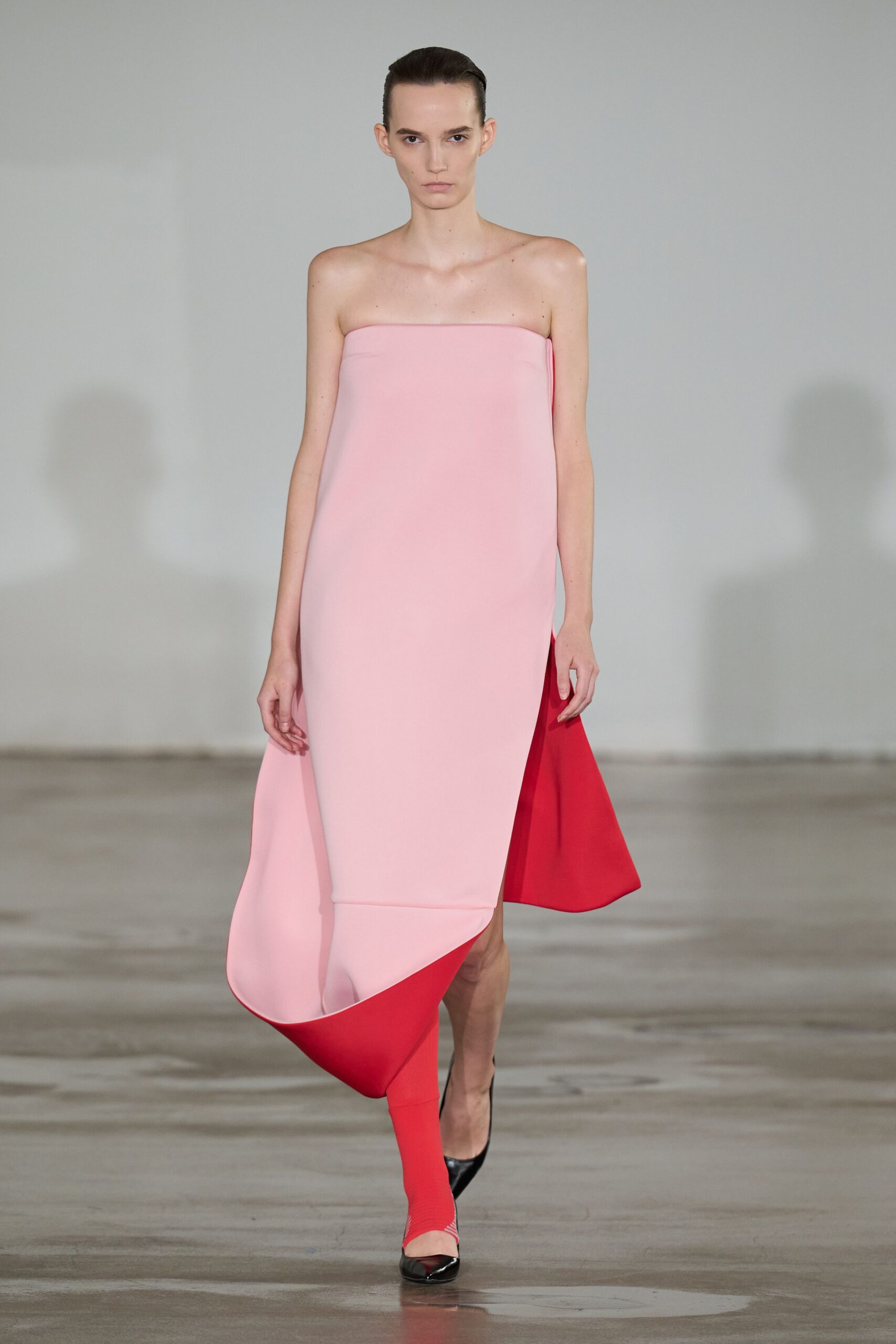Strapless pale pink column dress, foam-stiffened hem curling open to scarlet lining. Single tomato-red knit sock pulled to mid-calf. Black patent pointed pump with a narrow slingback strap. Crossing a museum atrium with a poster tube under one arm.