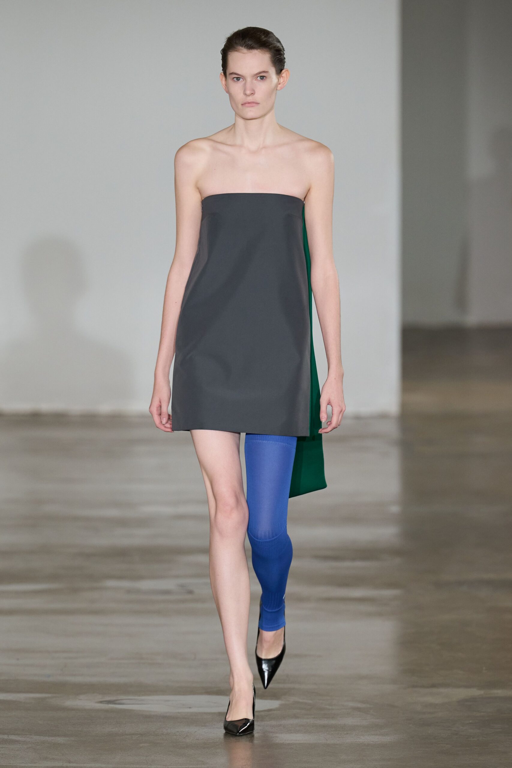 Charcoal shift cut in crisp satin, hem kicked slightly off square. Deep green tote hanging flat behind the hip. Cobalt ribbed legging worn on one leg only. Black patent pumps with narrow pointed toes. Walking across a near-empty office lobby before the lights fully switch on.