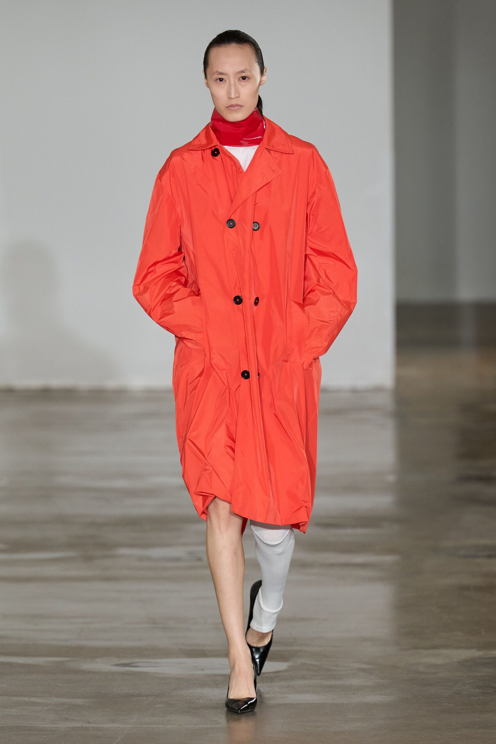 Glossy scarlet neck tube, creased where it folds under the chin. Fluorescent orange nylon trench, double-breasted with puckered sleeves and black buttons. Single white ribbed legging on one leg only. Black patent pumps with a sharp, pinched toe. Whatever you wear home when the rain starts before the car arrives.