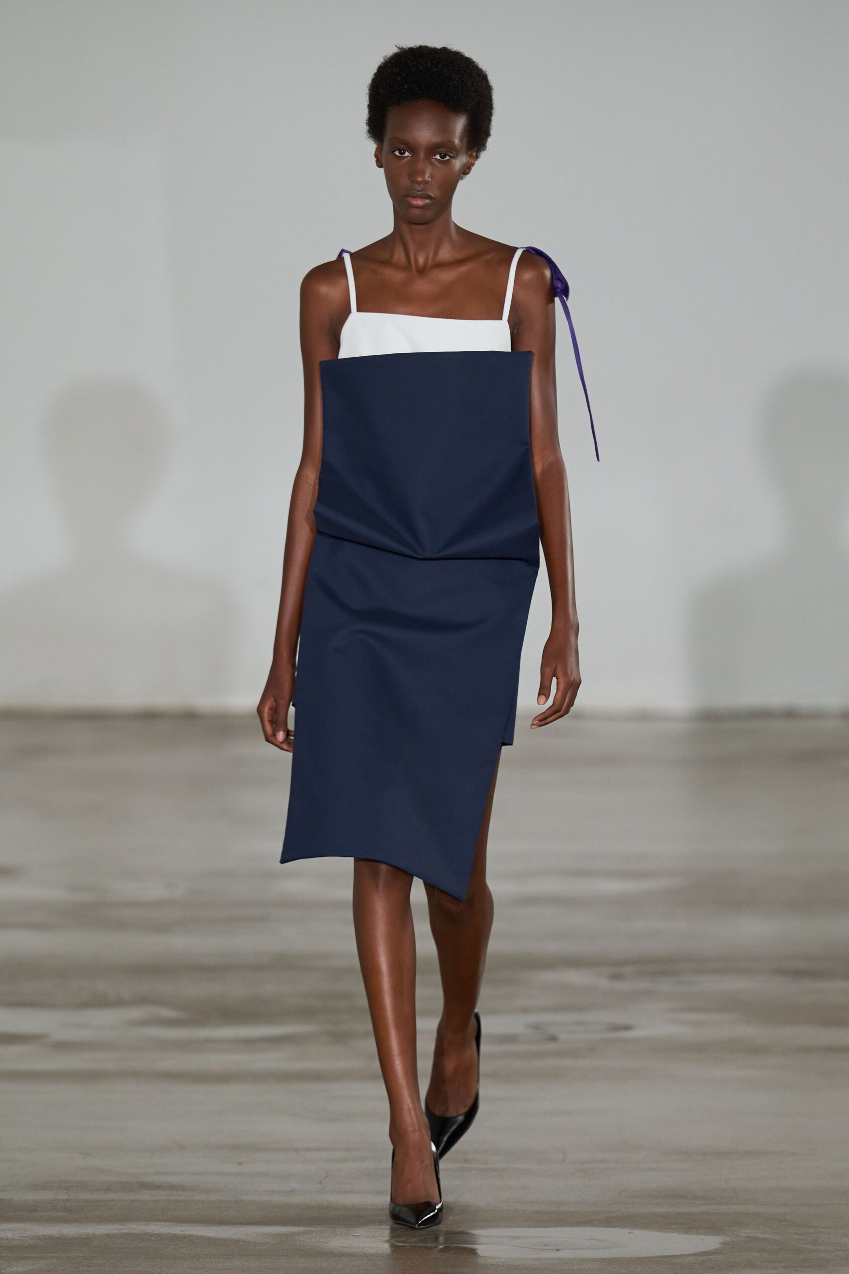 White camisole bodice meeting a navy folded panel that juts from the torso. Violet ribbon bow on one strap, one tail hanging past the arm. Black pointed pumps with a hard patent gleam. Walking across an emptied civic hall after the folding chairs are gone.