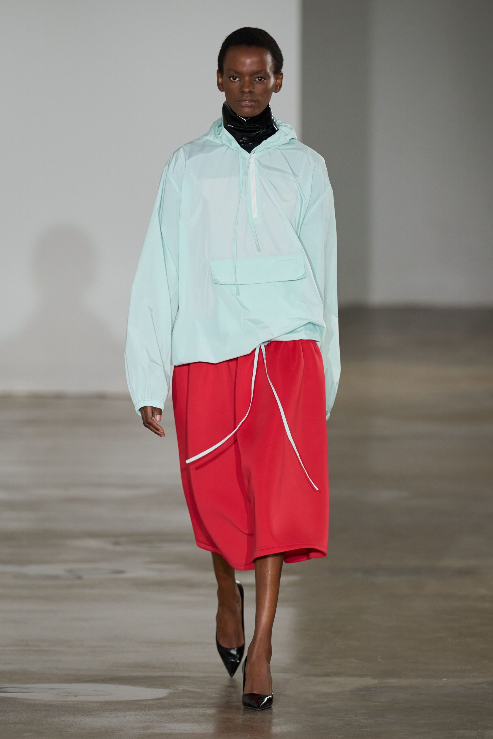 Glossy black vinyl tube hugging high under the jaw. Pale mint nylon anorak with half zip and skewed kangaroo pocket. Tomato red crepe culottes, white drawcord ends swinging past the knee. Black patent pumps, sharply pointed. Walking the seafront after rain, carrying lunch in the hood.
