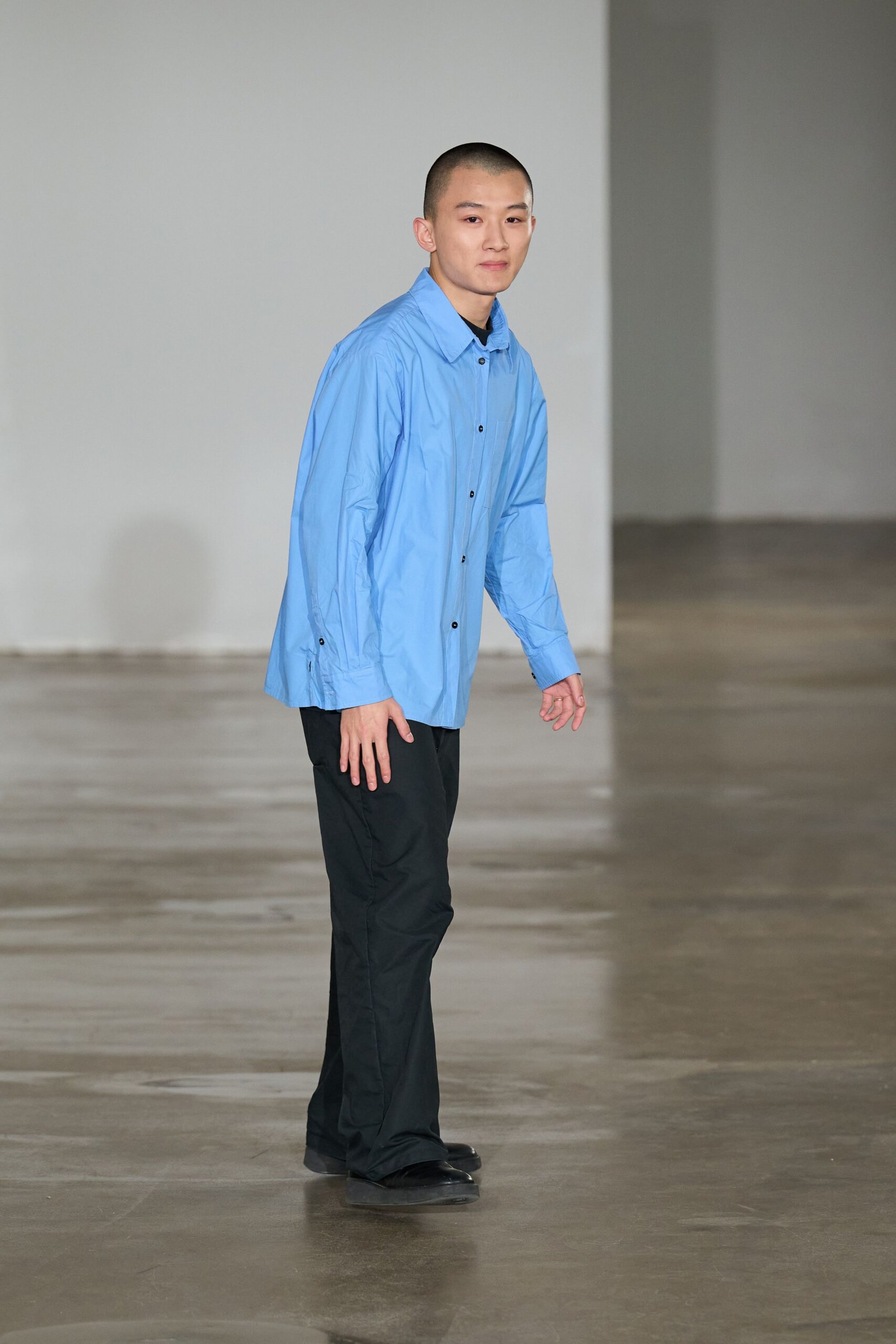 Pale blue poplin shirt hanging loose, black buttons punctuating the placket. Black crewneck just visible at the collar. Straight black trousers with a faint crease and slight break. Square-toed black leather shoes. Walking across an empty studio floor before the paint smell lifts.
