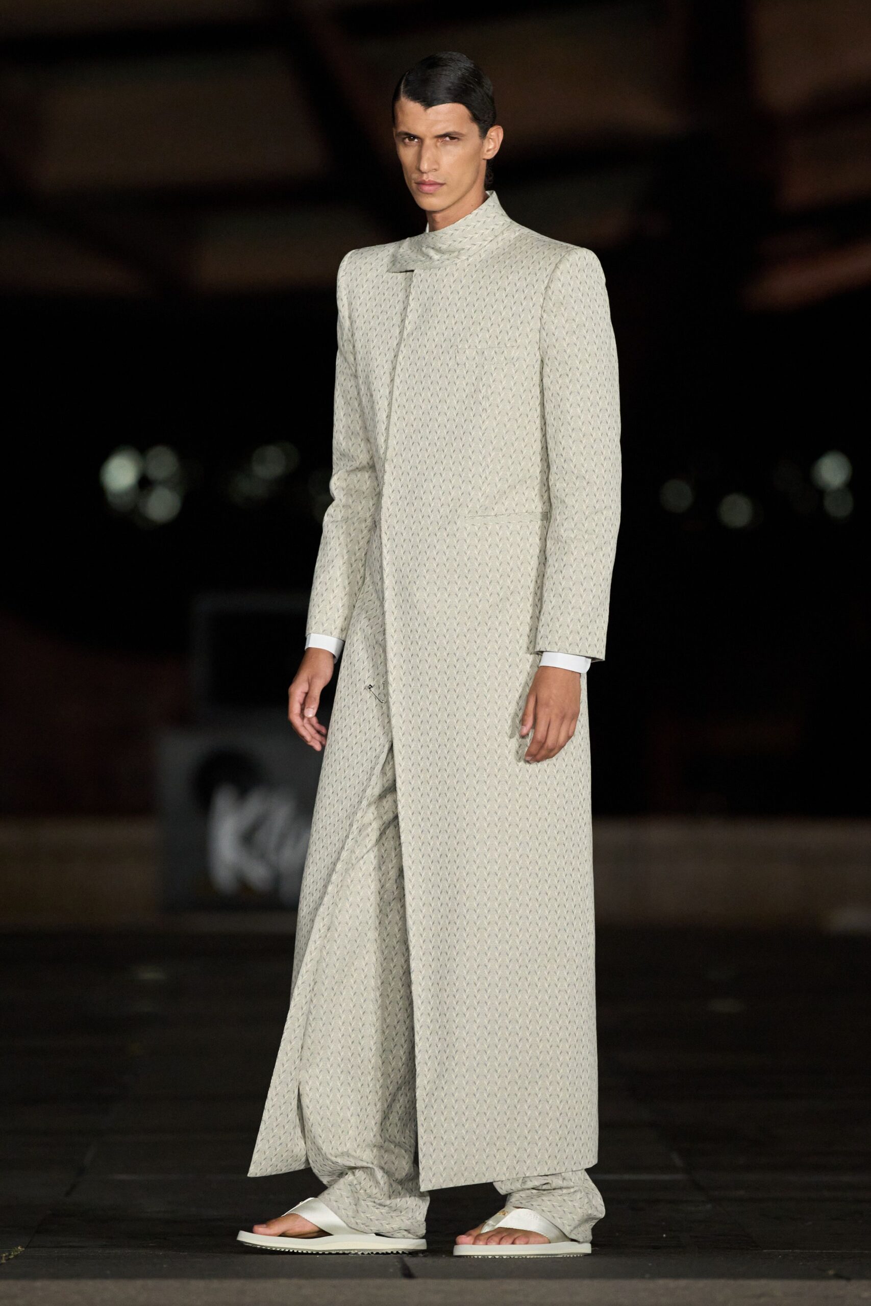 Long ivory coat in tiny chevron jacquard, cut with squared shoulders. White cuffs peeking cleanly below the sleeves. Matching wide trousers collapsing into soft folds over the feet. White thong sandals with thick straps and flat ridged soles. Temple courtyard after rain, moving carefully across the wet stone.