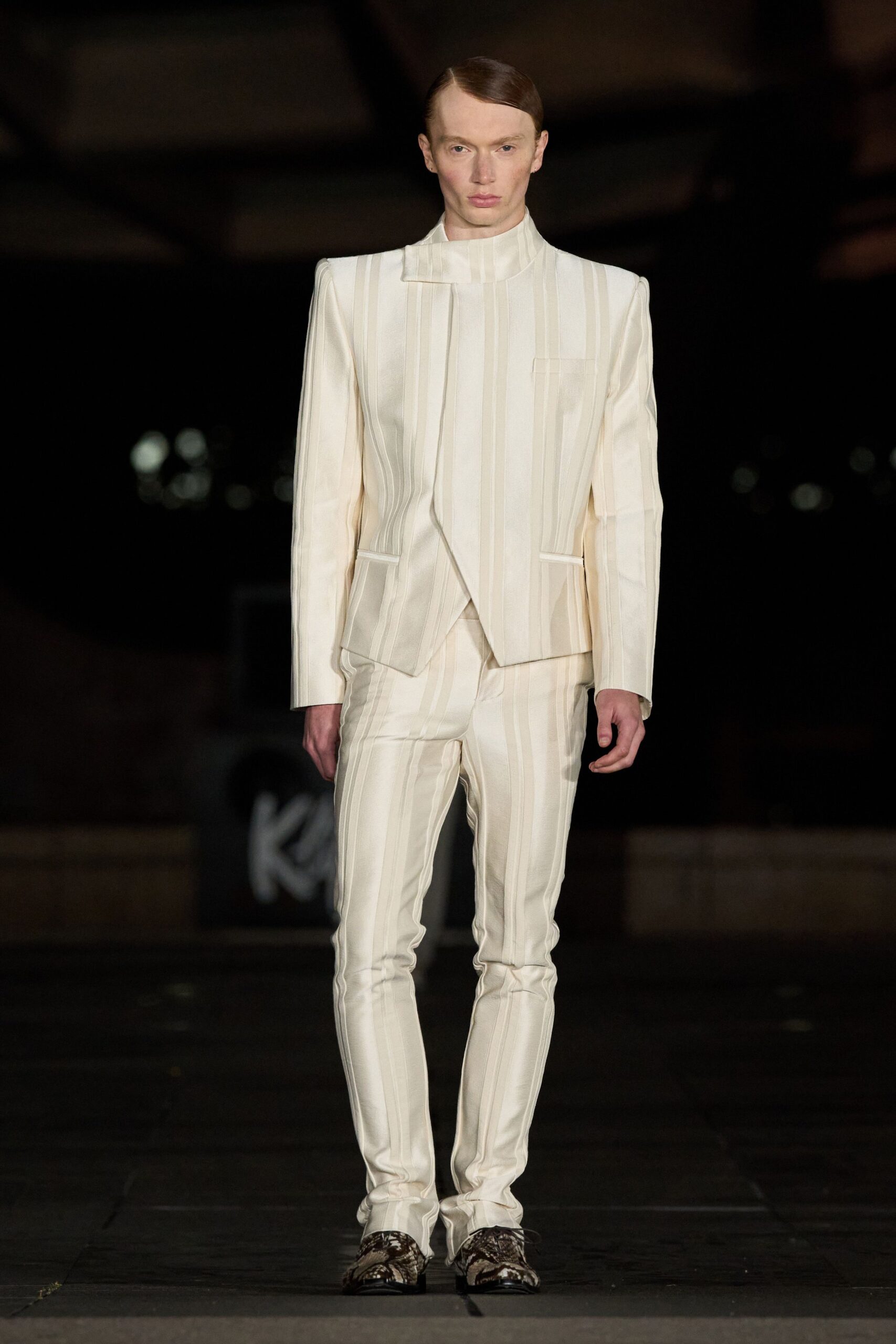 Ivory tailored jacket with raised vertical ribs and a folded stand collar. Matching slim trousers, the same ribbed stripes pulling into soft ankle stacks. Python-pattern derbies with blunt black soles. For crossing a hotel atrium at dawn before anyone else is dressed.