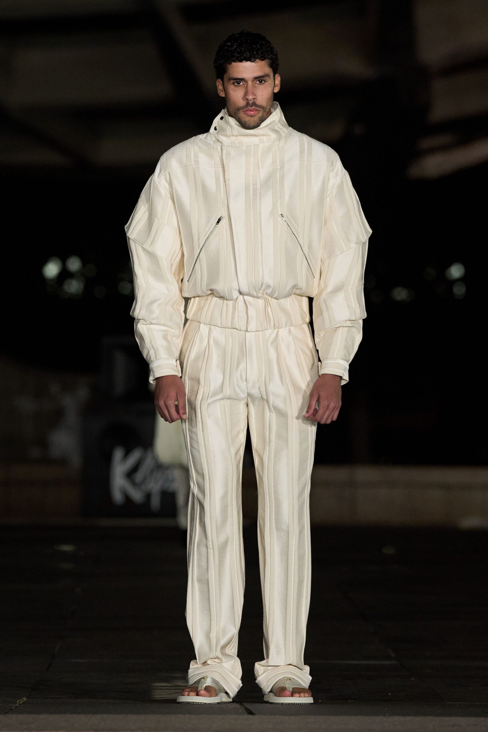 Ivory satin anorak with stitched vertical channels and diagonal zip pockets. Matching satin trousers, front seams falling into a soft break. White padded thong sandals. Walking out to a hotel pool before sunrise, towels still stacked.