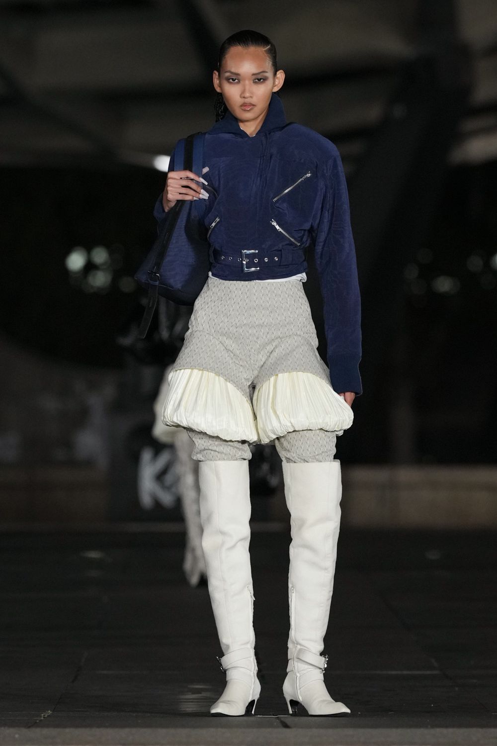 Ink velour hooded jacket with silver zips and a belted hem. Matching velour tote on a black webbing strap. Pale quilted trousers swelling into white ballooned pannier pads. White thigh-high boots with side zips and tiny metal buckles. Whatever you wear home when the taxi never came.