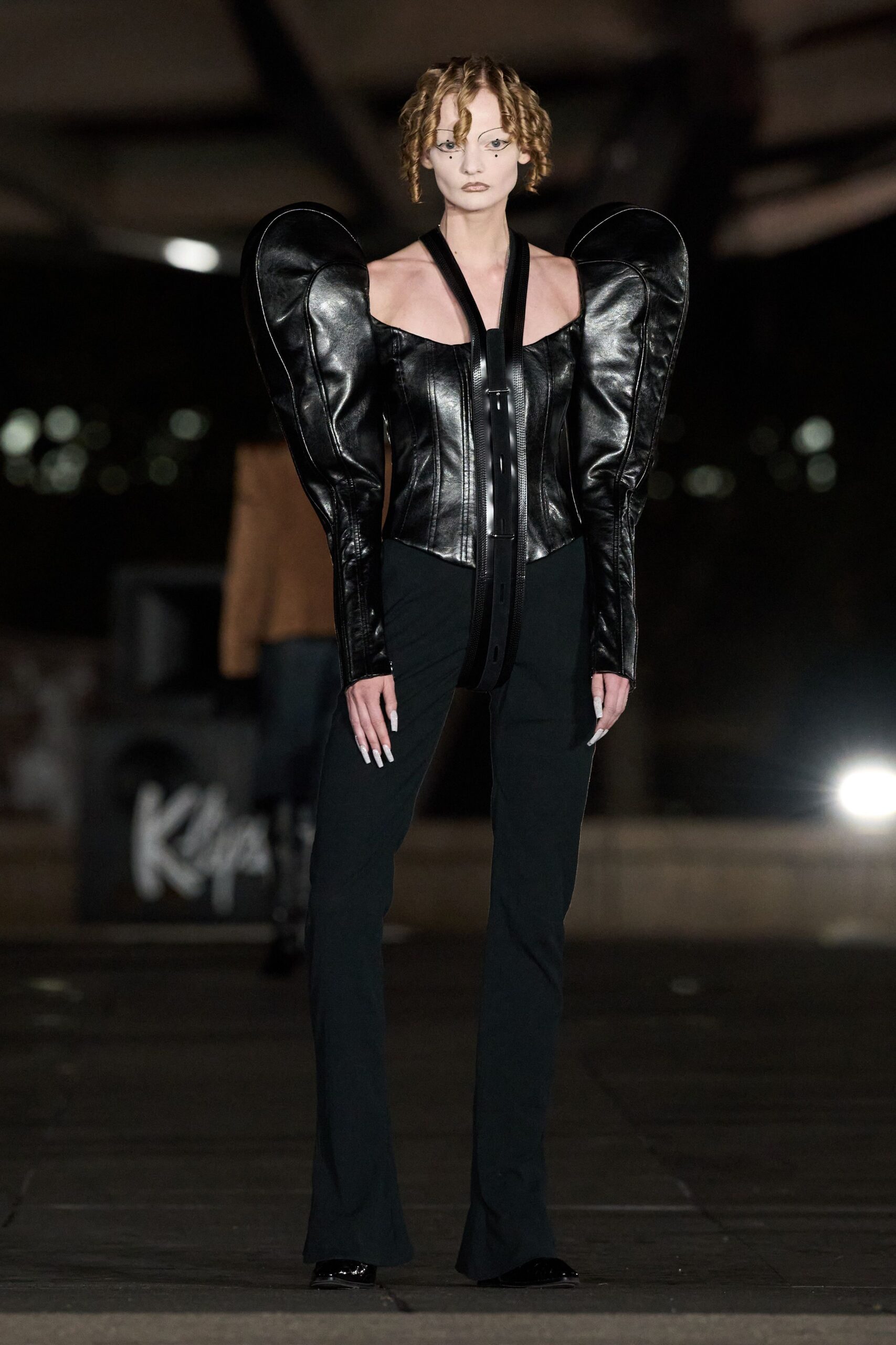 Black leather corset with towering crescent sleeves and a belt-like halter drop. Black narrow trousers breaking lightly over the shoe. Glossy black pointed toes. Under a highway overpass after midnight, concrete still breathing out heat.