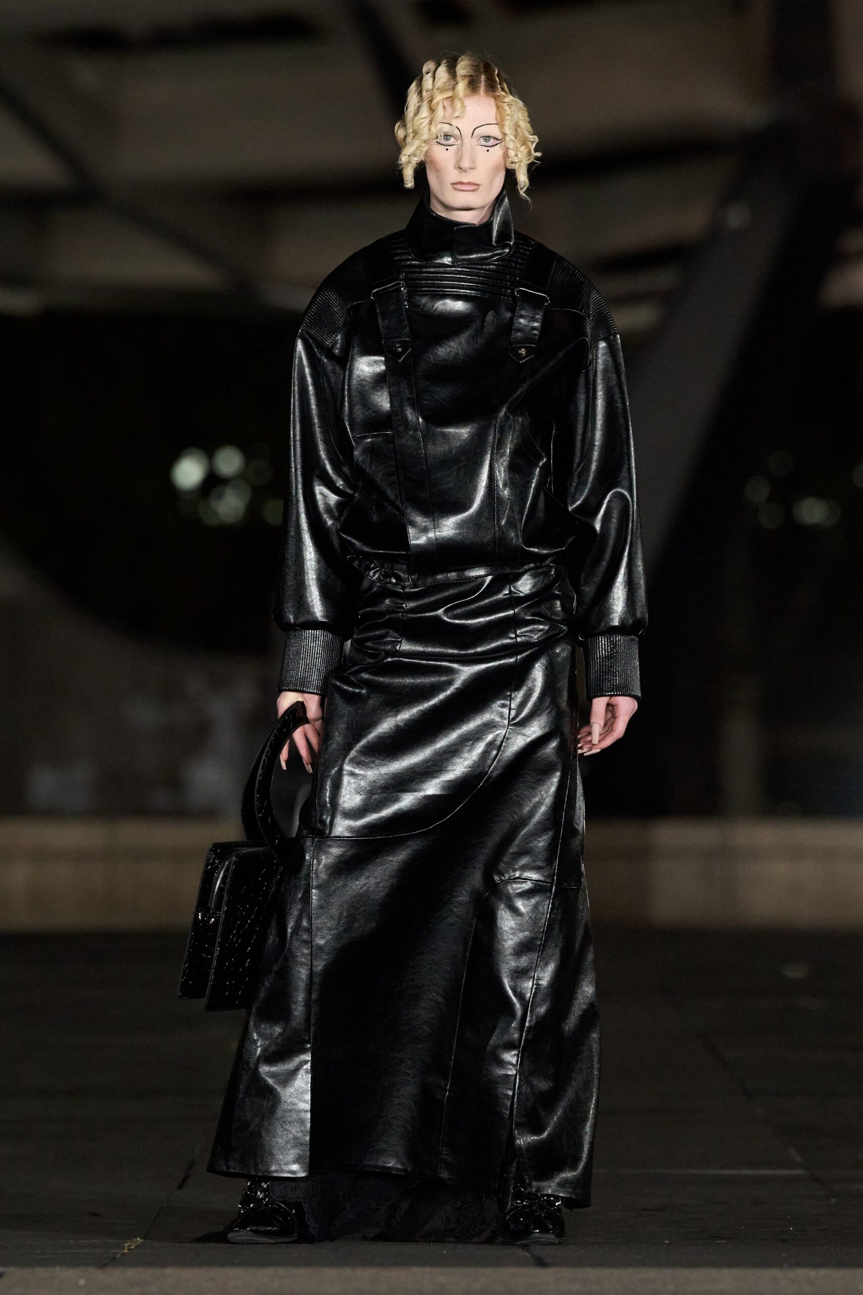 Black leather dress with a high funnel neck and ribbed shoulder panels. Wide charcoal knit cuffs cinching the sleeves at the wrist. Glossy black box bag punched with tiny regular studs. Black patent shoes barely visible under the dragging hem. Crossing an empty concourse after the last train, carrying nothing soft.