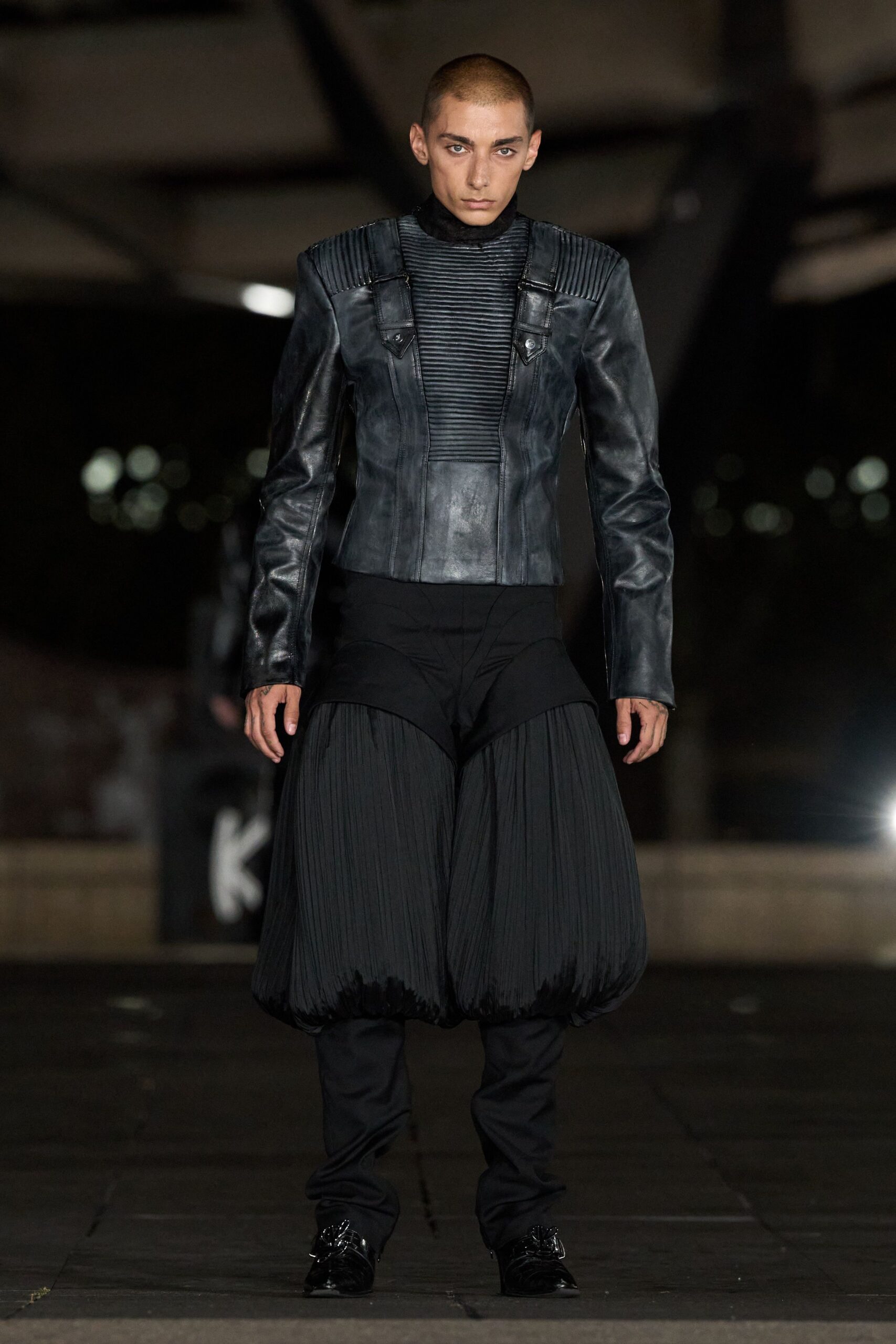 Black leather jacket with ribbed shoulder panels and one exposed snap. High-neck knit inset, tightly corded across the chest. Black trousers with pleated balloon thighs collapsing into narrow legs. Pointed black shoes with looped cord tassels at the vamp. Crossing an empty depot platform after the last train has gone.