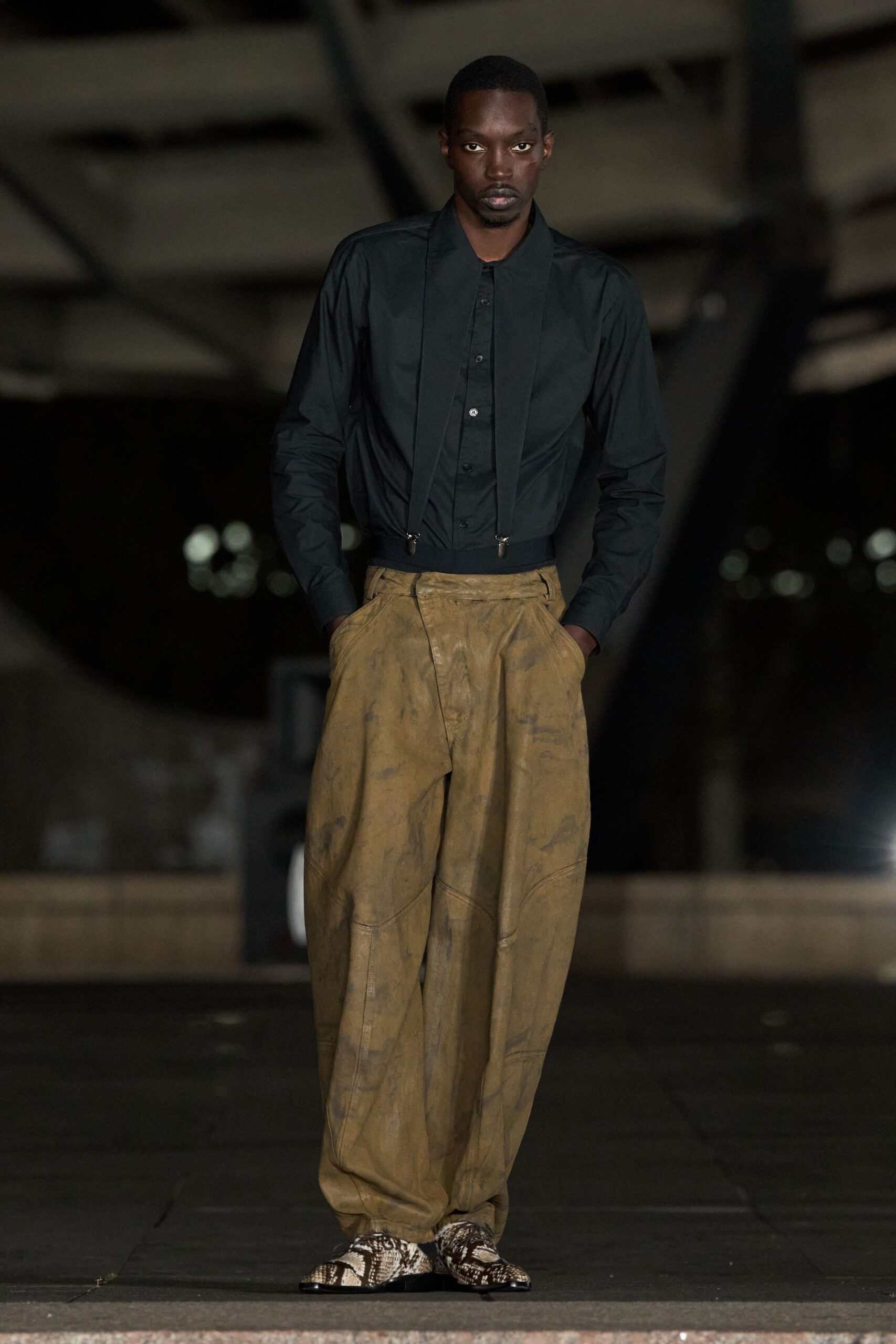 Black poplin shirt with narrow placket and long suspender straps clipped at the waist. Ochre leather balloon trousers, marbled with dark smudges and curved panel seams. Python-print pointed shoes. For crossing an empty loading dock after rain, still in work clothes.