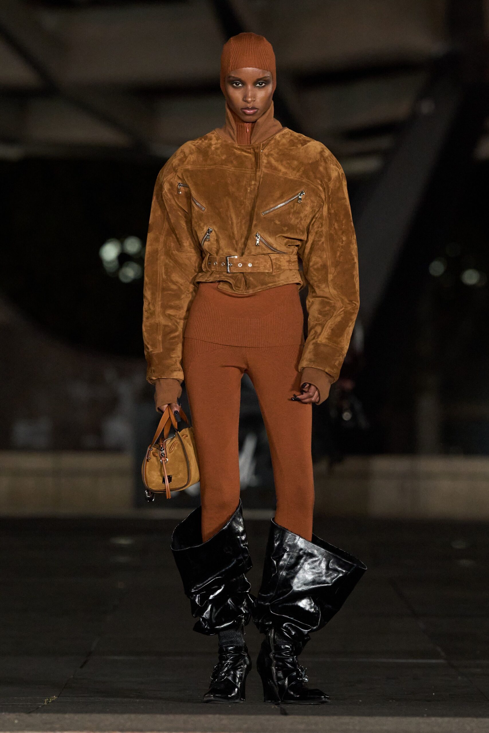 Rust rib knit balaclava zipped to the throat. Tobacco suede bomber, four diagonal zips and a buckled waist. Matching rib leggings with a wide folded waistband. Ochre suede bowling bag with long leather zip pulls. Black patent boots collapsing into oversized slouched funnels. Crossing an empty concourse after the last train left.