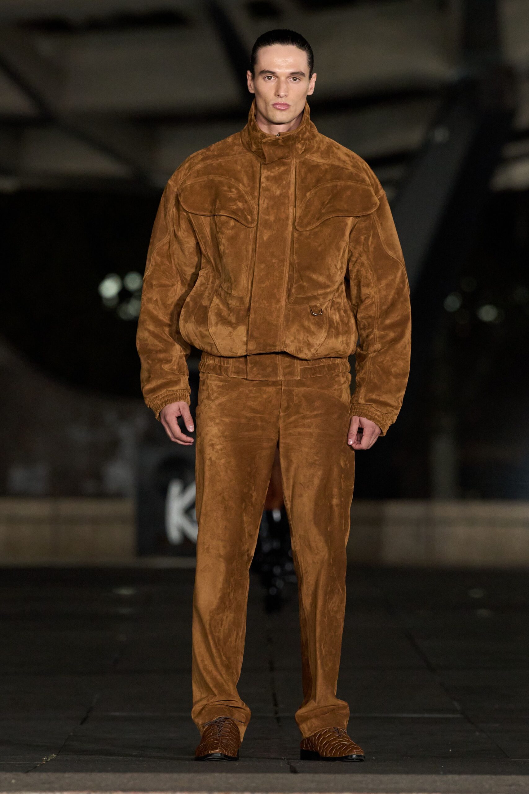 Cinnamon suede blouson with a stand collar and curved flap panels. Matching suede trousers, softly crushed through the leg and pooling lightly. Chestnut woven leather square-toes with black stacked soles. For crossing an empty airstrip just after the rain stopped.