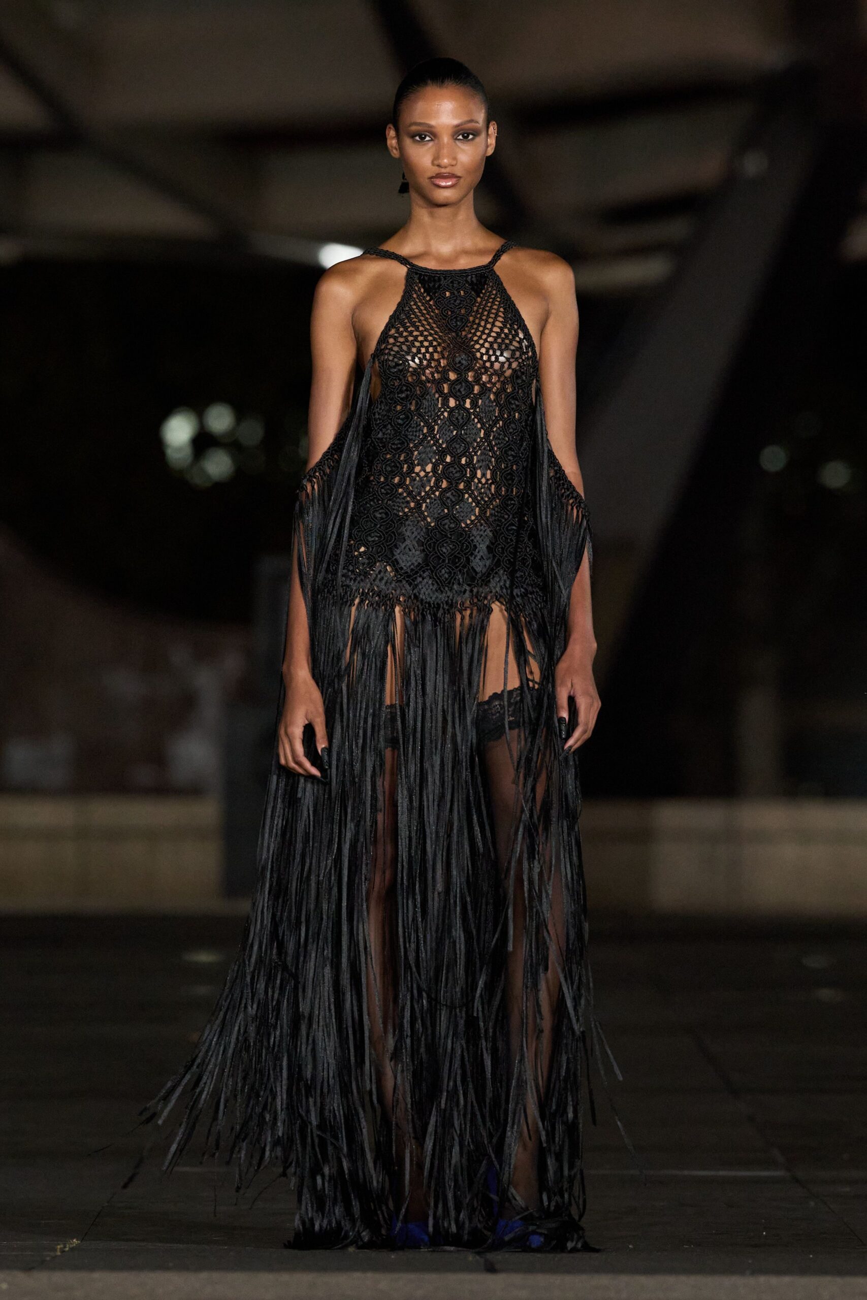Black macrame halter dress, openwork medallions dropping into ankle-grazing fringe. High-cut black briefs visible beneath the netted skirt. Sheer black thigh-highs with lace tops peeking through. Cobalt pointed heels flashing under the swinging fringe. Walking across wet paving after midnight, hem drinking up the puddles.
