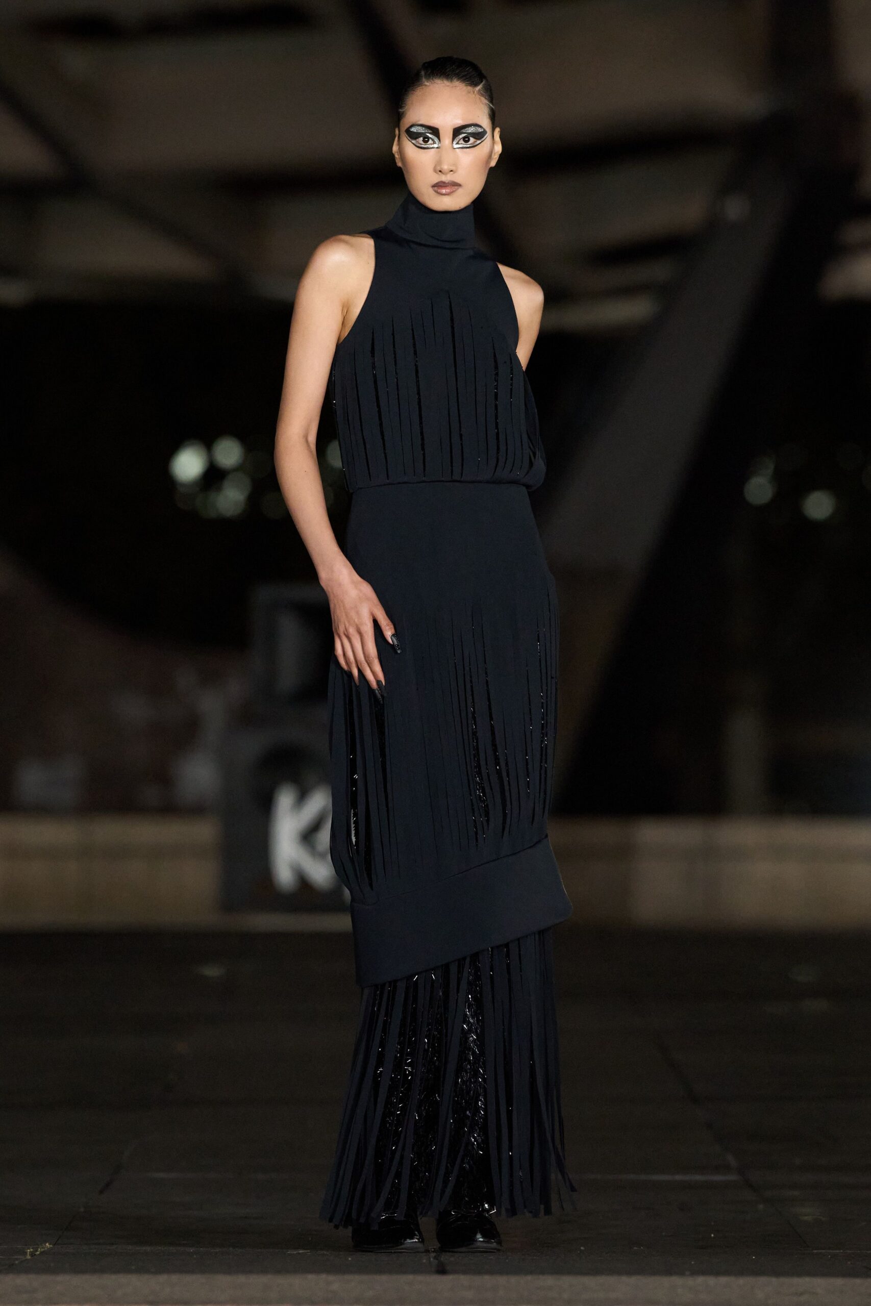 Black sleeveless column dress with a tall funnel neck. Narrow matte strips falling in vertical panels, glossy underlayer flashing through. Black pointed boots barely visible beneath the moving hem. Crossing a museum atrium after dark while the fountains are turned off.