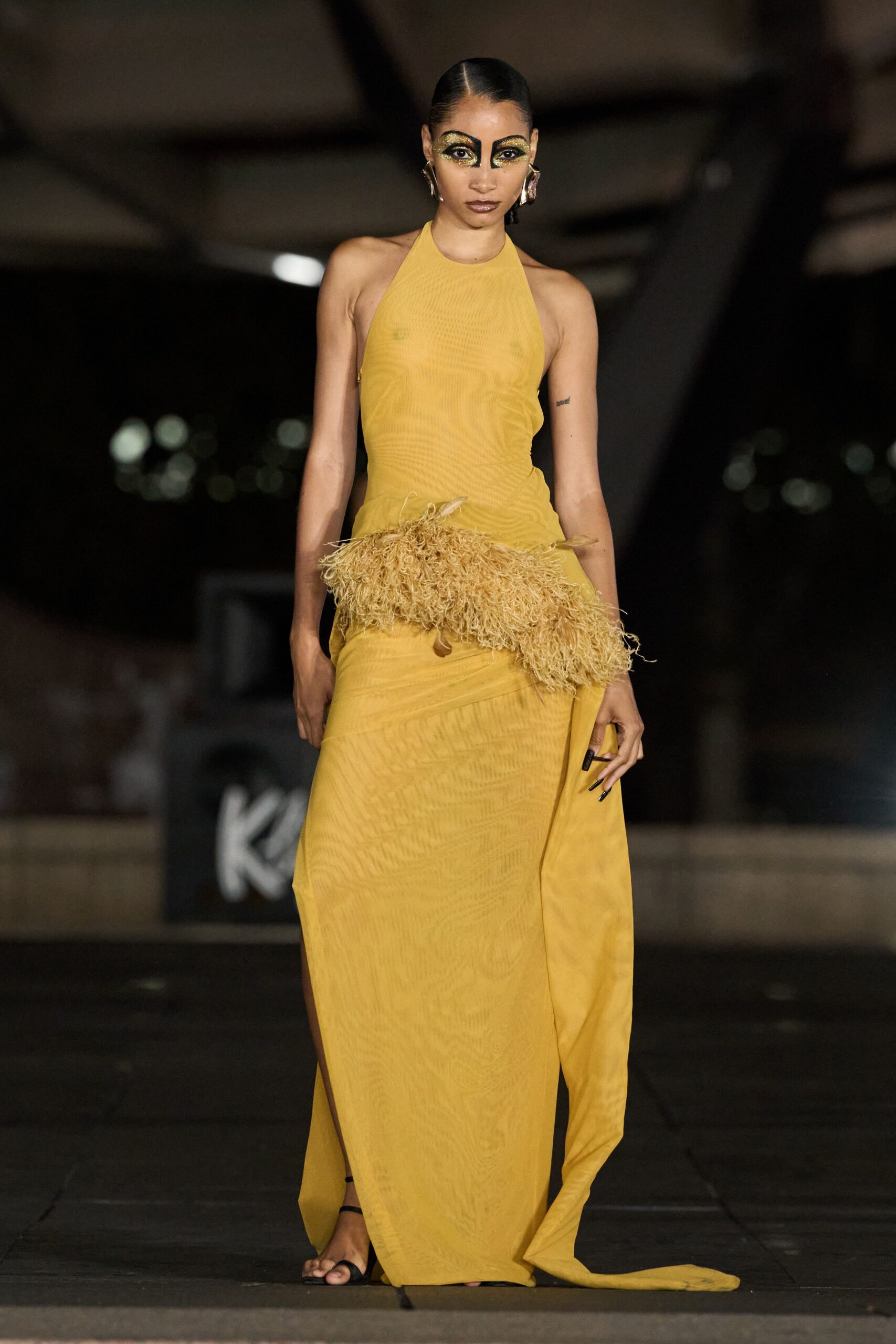 Mustard halter gown in sheer crinkled mesh, wrapped low with a trailing panel. Shaggy raffia bustle jutting from the hips, a few feather tips poking through. Black thong sandals, nearly bare against the hem. Crossing a hotel courtyard after midnight with pool water still on the tiles.