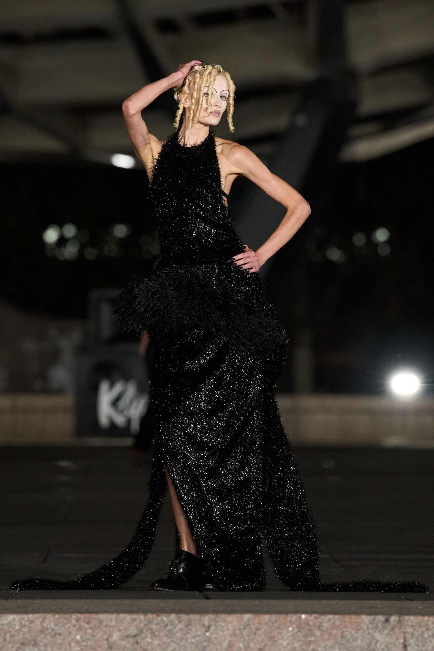Black halter gown in glittering eyelash knit, with a shaggy hip ruffle and dragging train. Square-toe black shoes, one vamp catching the light. After midnight on a station concourse, waiting for nobody now.