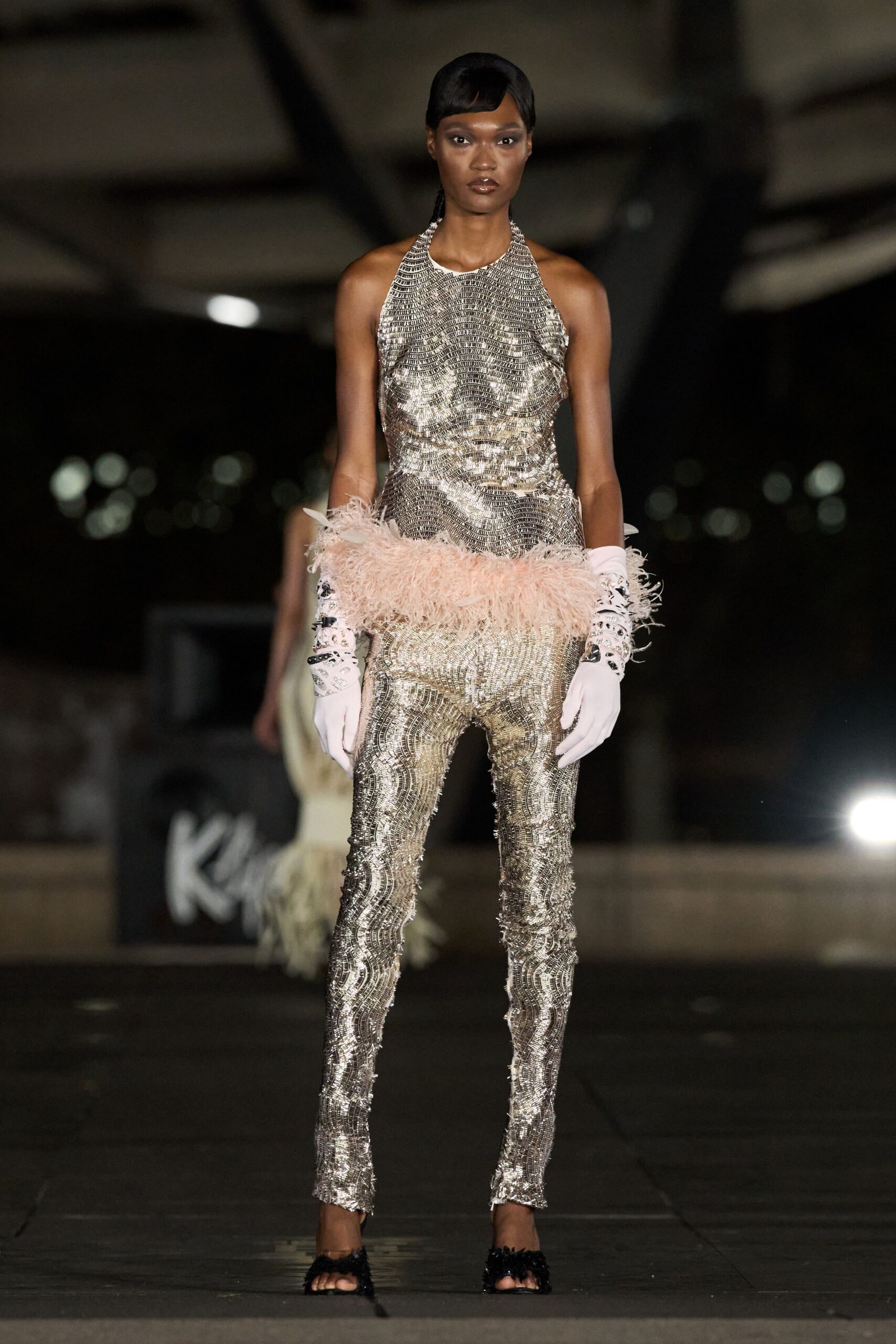 Silver paillette mesh in rippling bands, cut bare through the shoulders. Powder-pink ostrich fluff slung low across the hips. White opera gloves with crystal-studded black appliqué at the cuffs. Black feather-trimmed mules, toes left exposed. For the hotel ballroom after midnight, when the band is packing up.