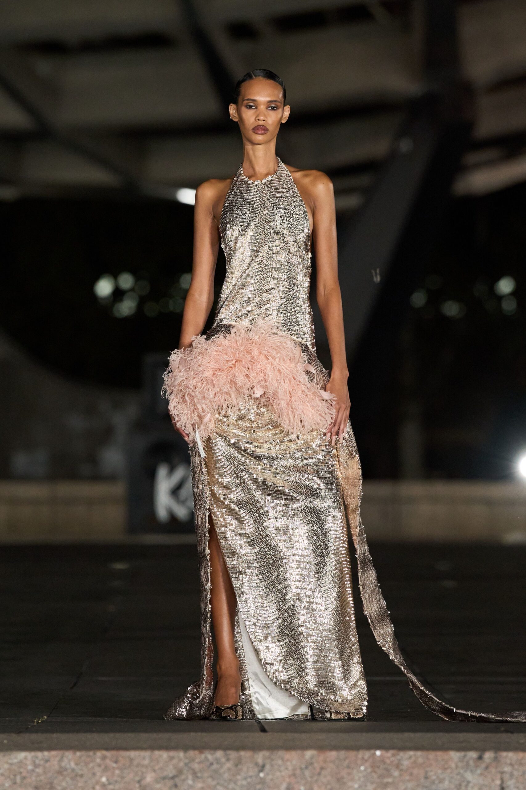 Silver paillette halter gown in rippling chevrons, slit high and train fraying long. Powder-pink ostrich feather boa bunched thick across the hips. Snake-pattern pumps peeking from beneath the hem. Crossing a hotel ballroom after midnight with petals ground into the carpet.