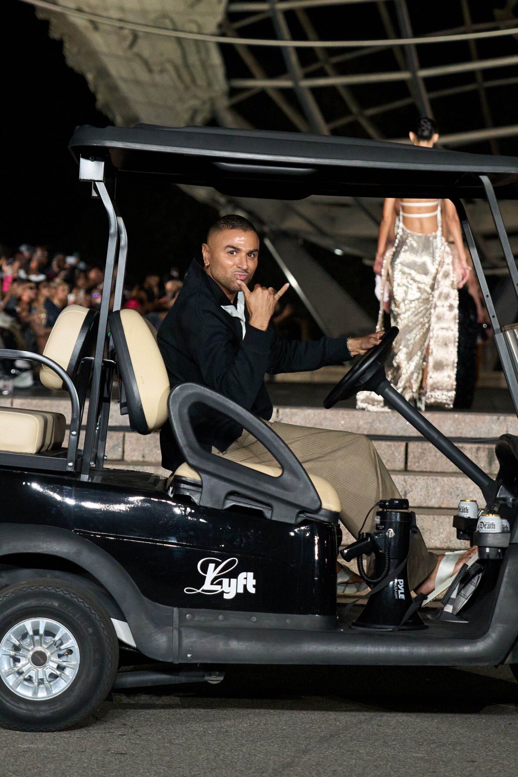 Black zip-front jacket with a crisp white insert peeking at the throat. Loose beige pinstripe trousers breaking softly over the footrest. White slide sandals with broad padded straps. For ferrying people between marble steps and the museum lawn after dark.