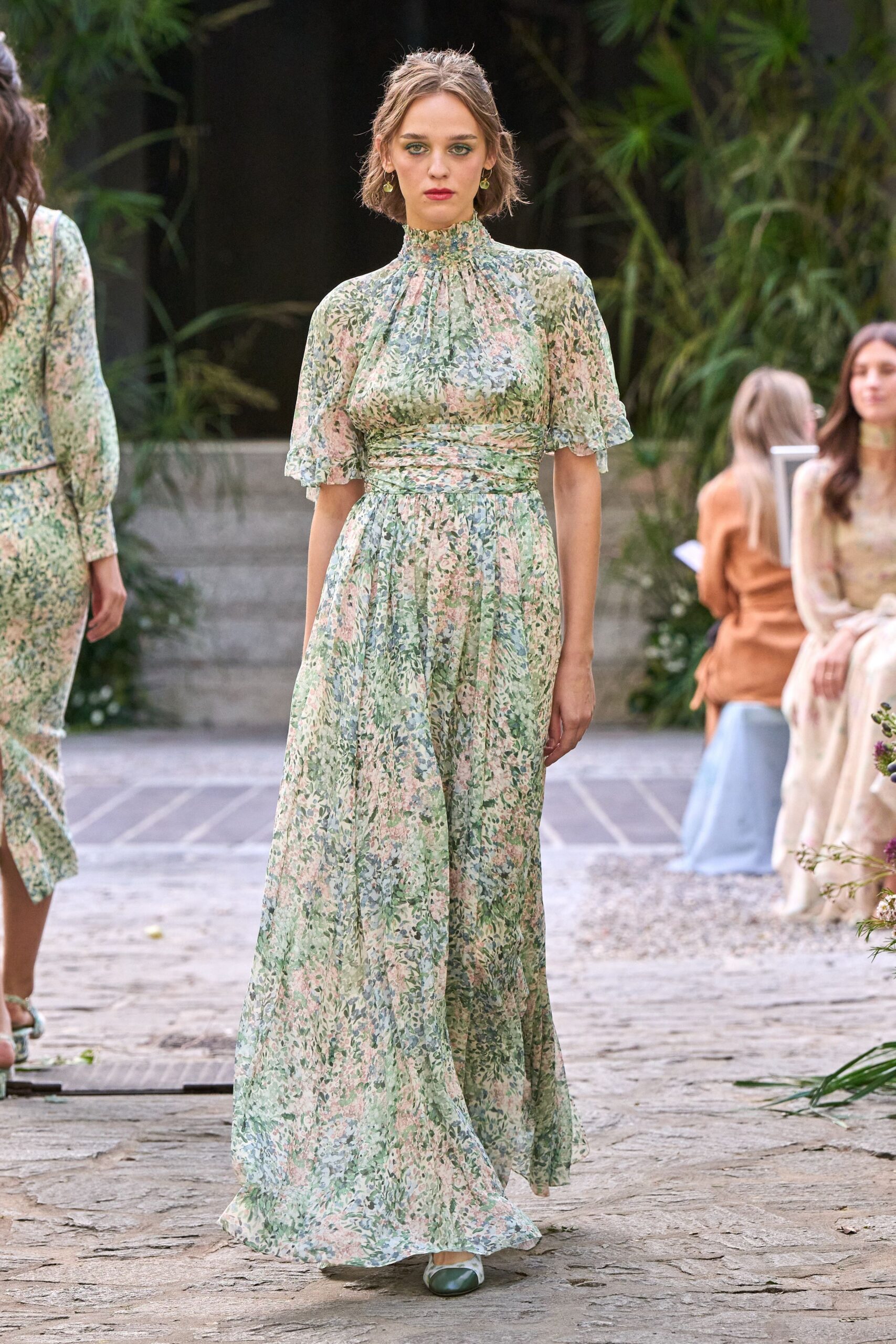 Sheer floral chiffon maxi dress, shirred high neck and ruched waistband. Pale green flats with darker capped toes peeking beneath the hem. Small drop earrings. Garden wedding spillover, walking back across wet stone after the vows.