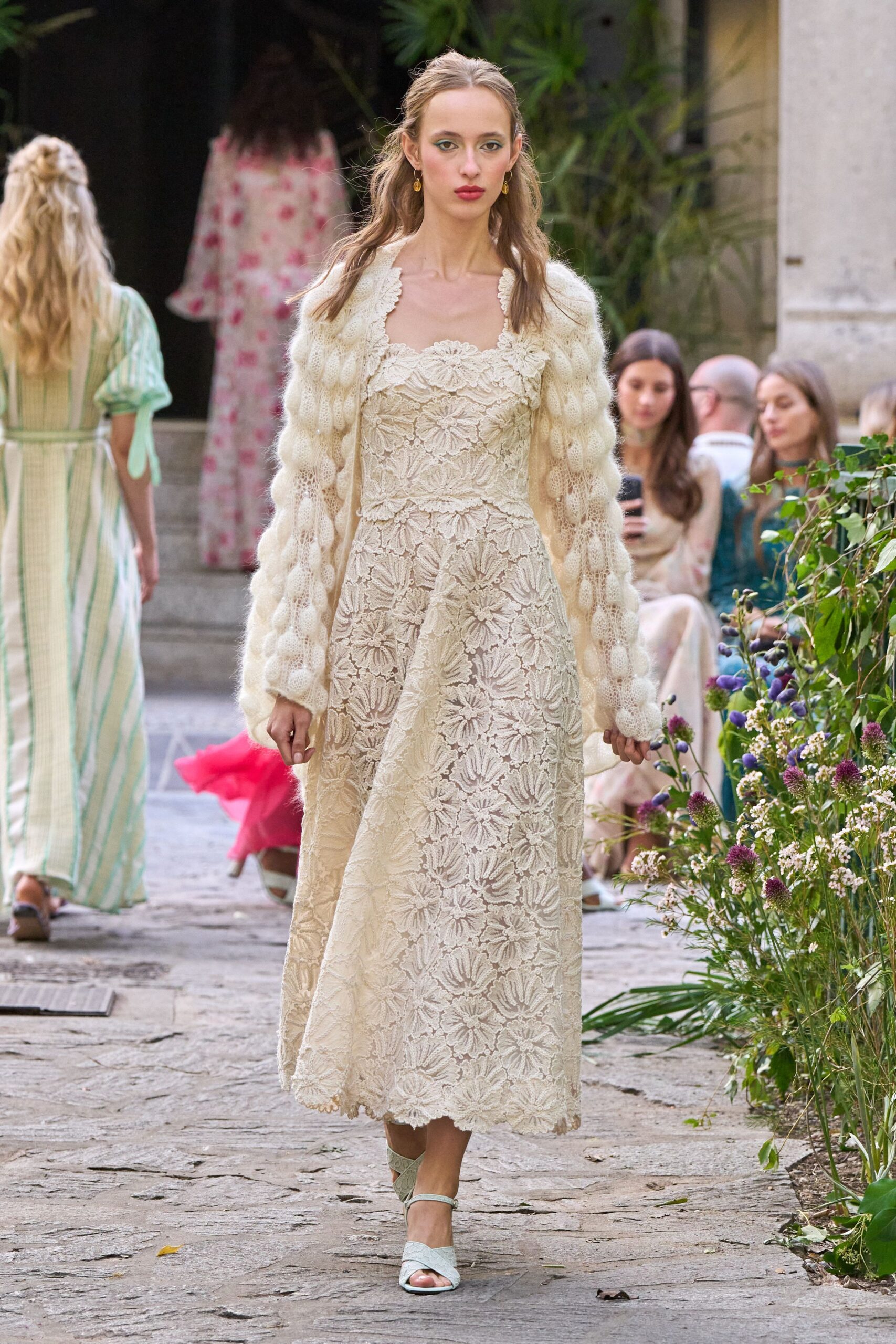 Cream mohair cardigan with puffed cable sleeves hanging past the hands. Ivory lace dress with dense daisy motifs and a scalloped square neckline. Pale ankle-strap sandals with softly quilted crossover bands. Walking through a monastery garden just after the paving stones have dried.