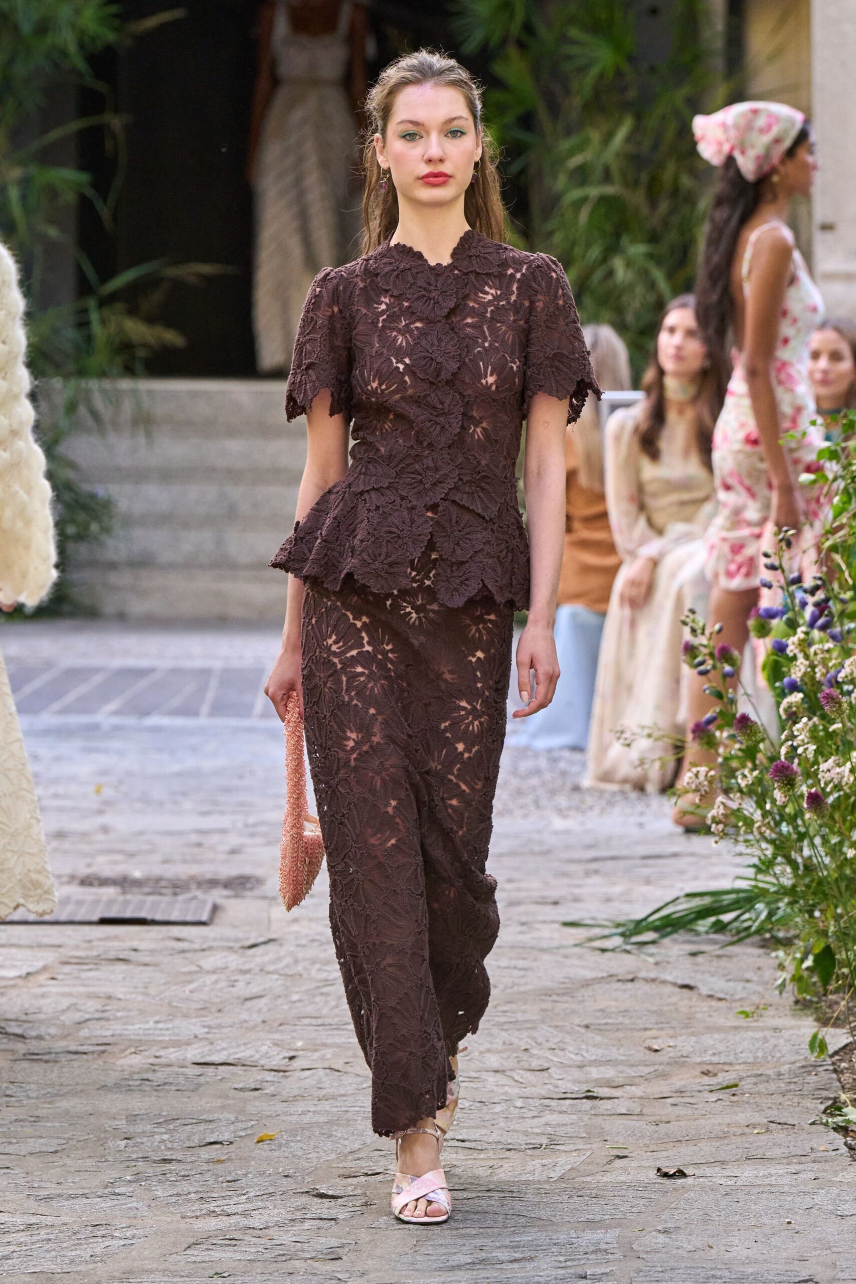 Espresso floral lace top over nude lining, petals scalloping the peplum. Matching column skirt in the same raised lace, hem grazing the instep. Blush beaded pouch dangling from the hand. Pale pink satin sandals with narrow crossover straps. Garden wedding reception, the air still damp after afternoon rain.