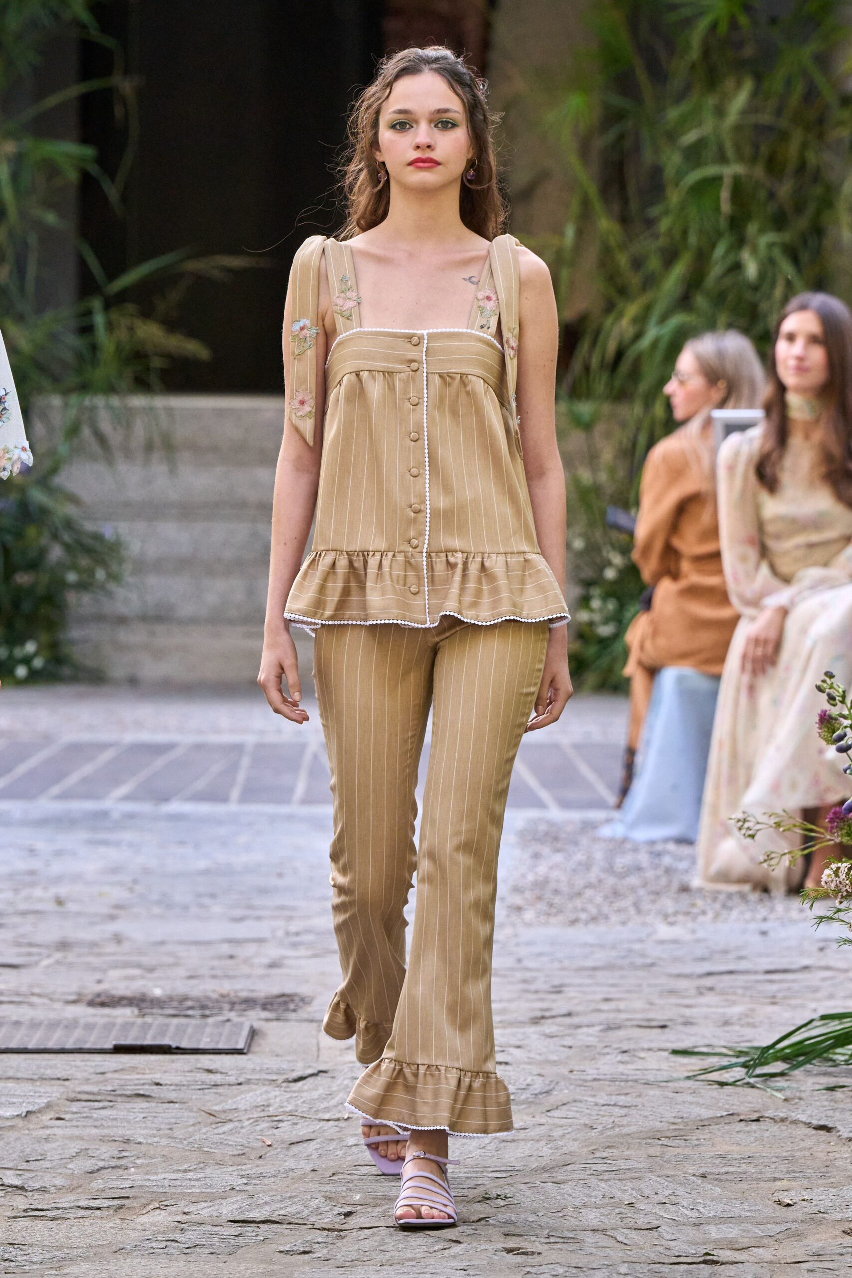 Camel pinstriped babydoll top with pearl picot edging and floral straps. Matching slim trousers, tiny ruffle hems brushing over the foot. Lilac lace-up sandals with squared toes. Garden wedding rehearsal, humidity still clinging to the stone path.