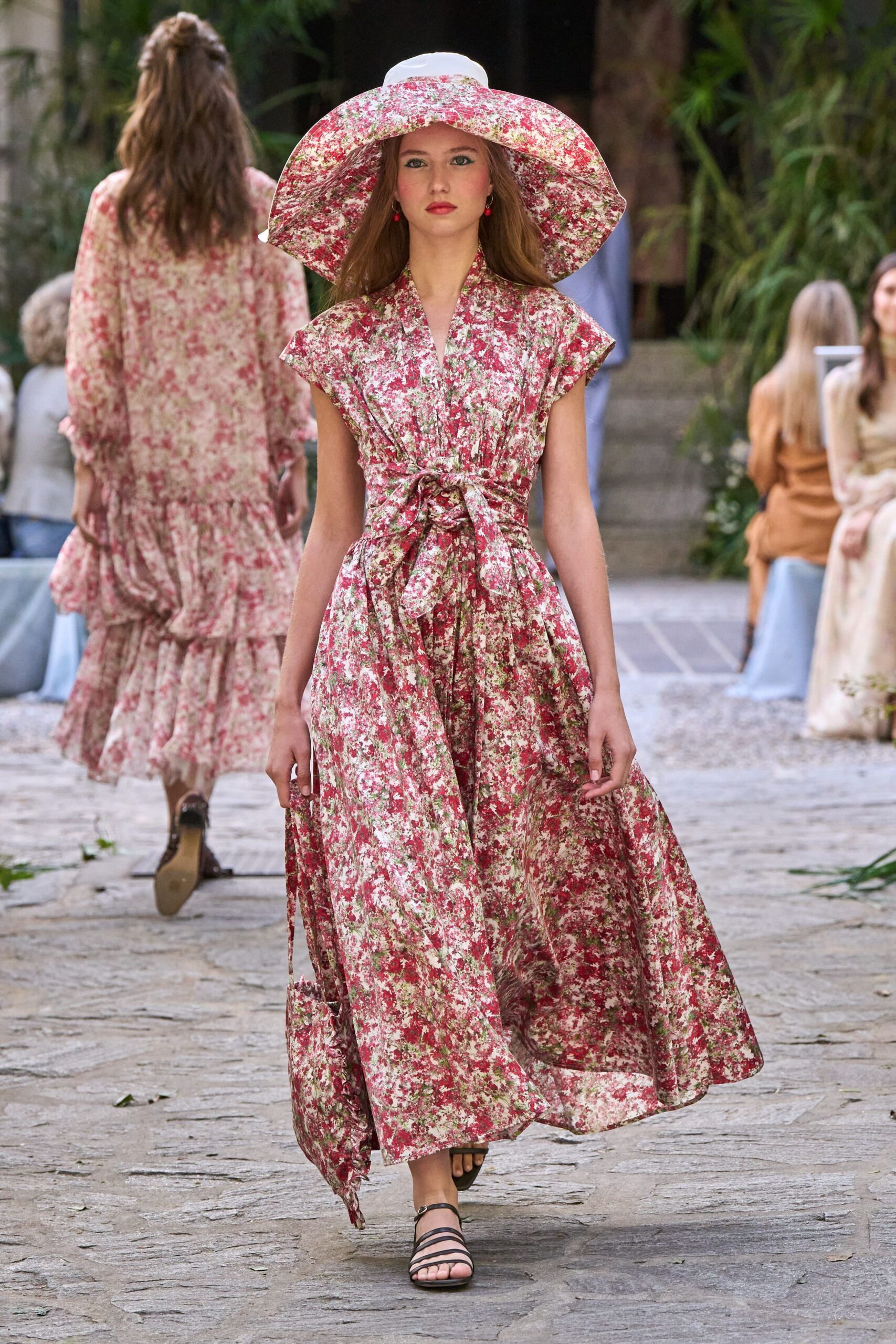 Oversized brim in matching pink ditsy floral cotton. Small cherry-red drop beads. Pink and moss floral wrap dress, cap sleeves and a bulky tied waist. Matching floral tote dangling from one hand. Flat black sandals with slim crossing straps. Walking back from a garden wedding after the sun has dropped.