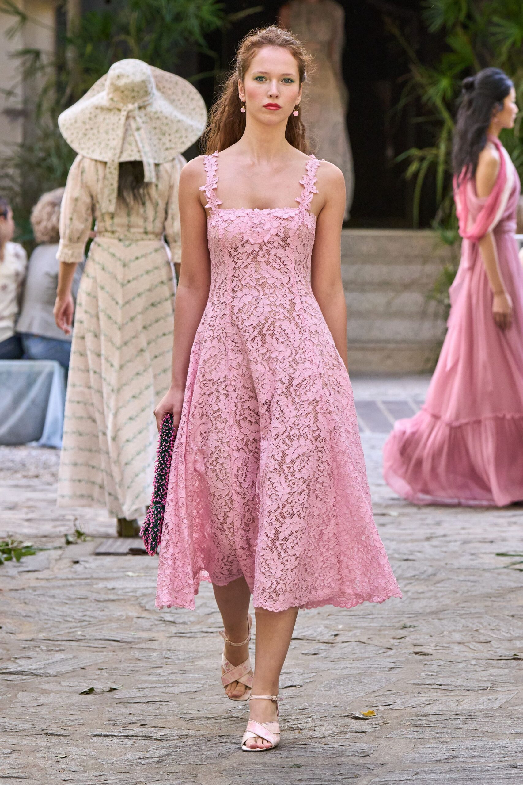 Blush floral lace dress with scalloped straps and a full tea-length swing. Tiny pink drop earrings. Beaded pouch crowded with pink and black fringe. Pale ankle-strap sandals with crossed satin bands. Garden wedding reception, just before the cake is carried out.