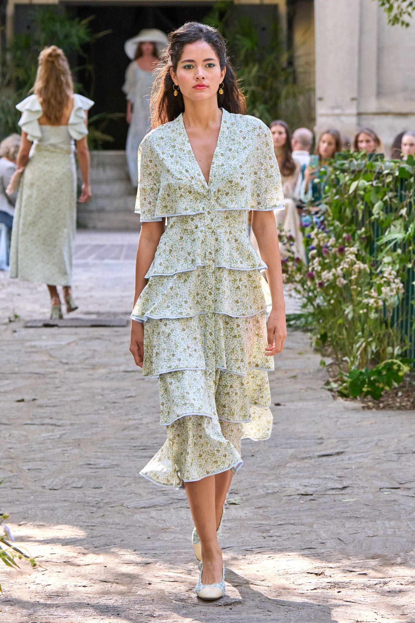 Ivory floral midi dress with cape sleeves and tiered ruffles edged in white piping. Small gold drop earrings. Cream cap-toe pumps in matching floral fabric. Garden wedding aisle, the hour when the stone still holds afternoon heat.