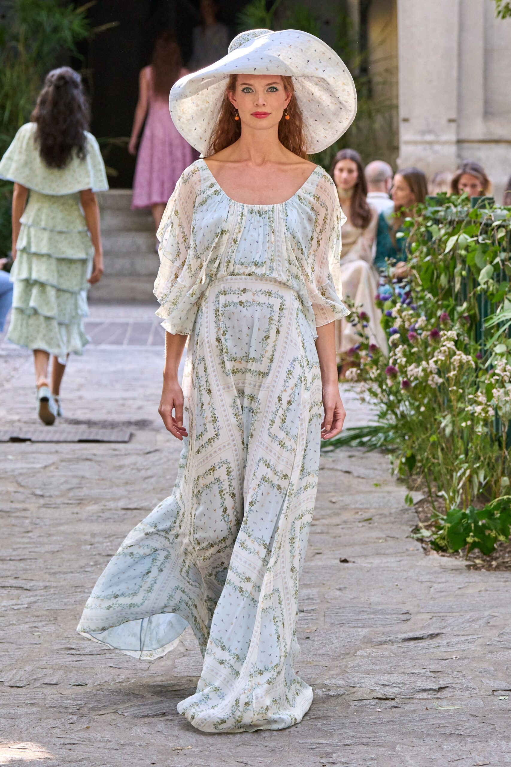 Oversized white sunhat, brim collapsing in a soft uneven wave. Tiny orange drop earrings. Powder-blue floral chiffon dress with a gathered square neckline and drifting hem. Walking the garden aisle before the chairs are filled.