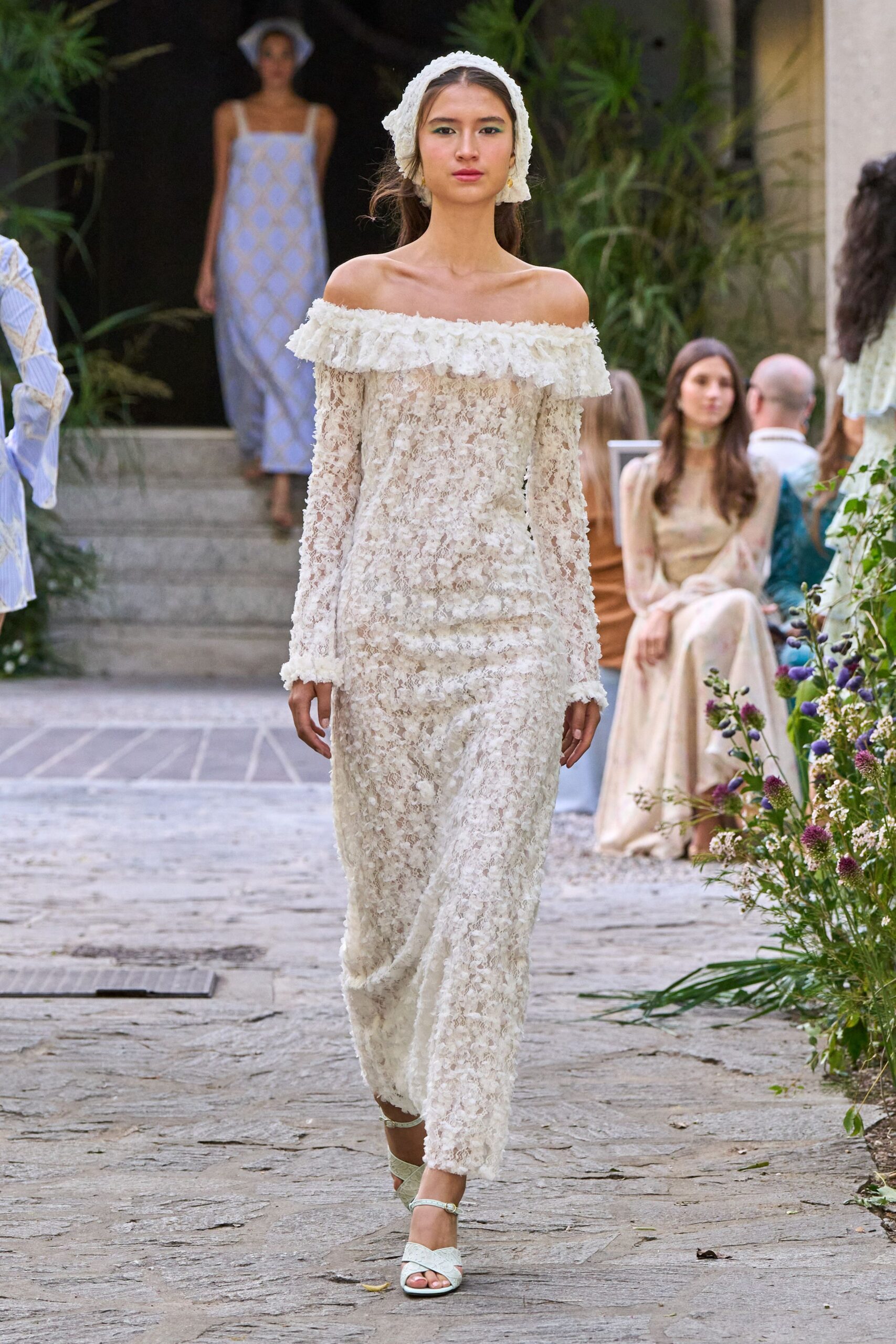 White lace headscarf tied low, scalloped edge framing the ears. Off-shoulder ivory lace column dress with a ruffled neckline and petal-thick surface. Pale ankle-strap sandals with quilted crisscross bands. Garden wedding, the stone path still damp from morning watering.