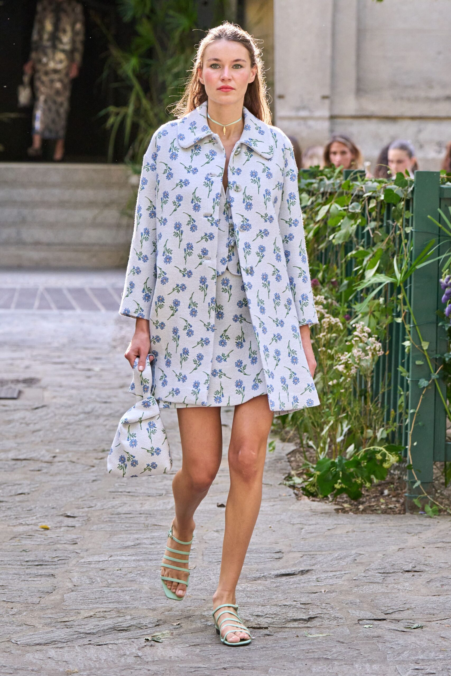Pale textured coat scattered with blue sprig florals and a rounded collar. Matching mini dress peeking through, tied at the bust. Mint cord choker with a single shell drop. Soft floral pouch bag, slouched in the hand. Mint strappy flat sandals. Garden ceremony spillover, someone carries cake plates back through the side path.
