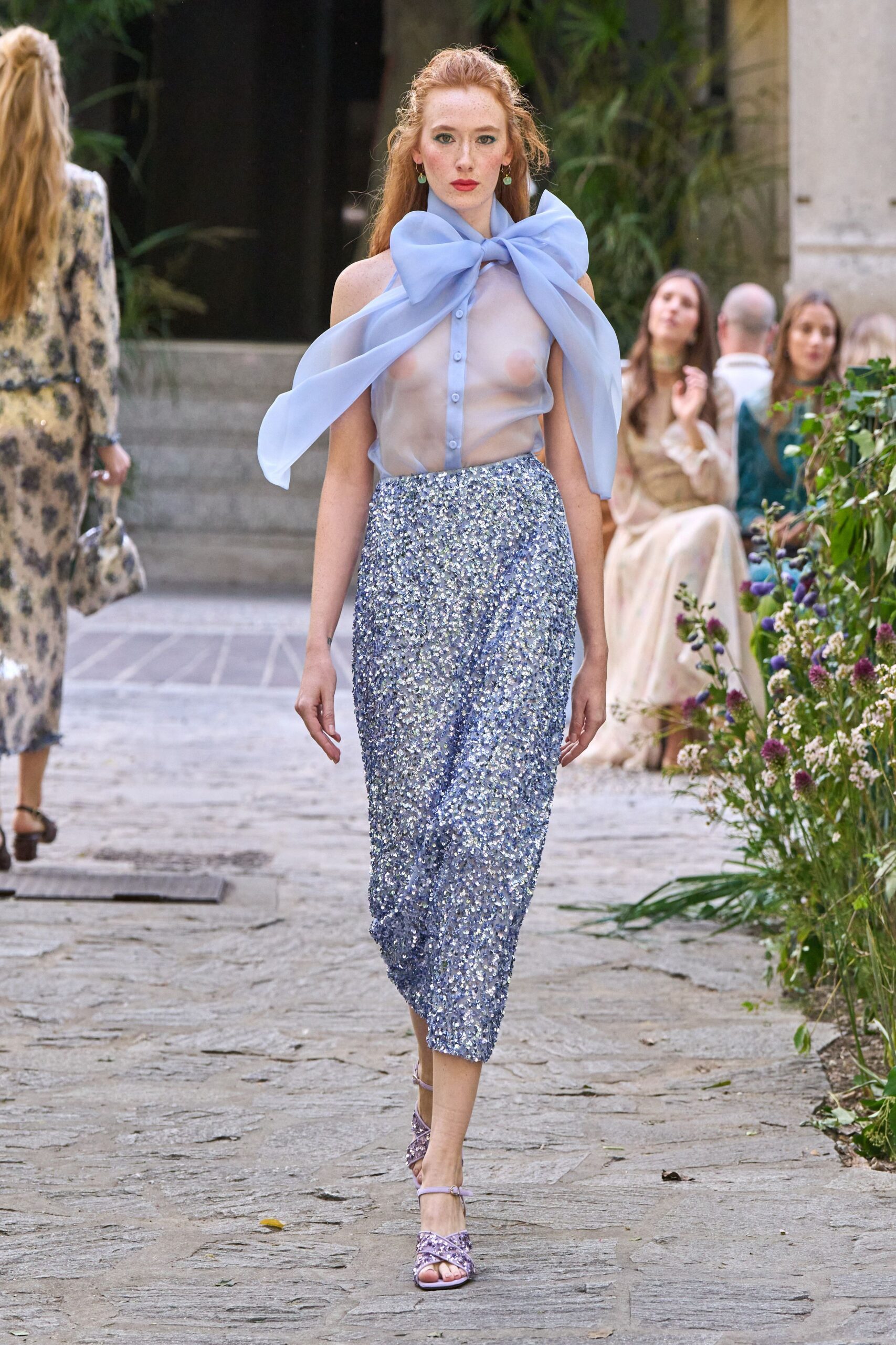 Sheer powder-blue organza blouse with a huge bow knotted at the throat. Pencil skirt packed with pale lilac paillettes, tapering tight at the calf. Lilac ankle-strap sandals with crossed glitter bands over the toes. Small mint drop earrings. Garden wedding aisle, the hour when the champagne has started to work.