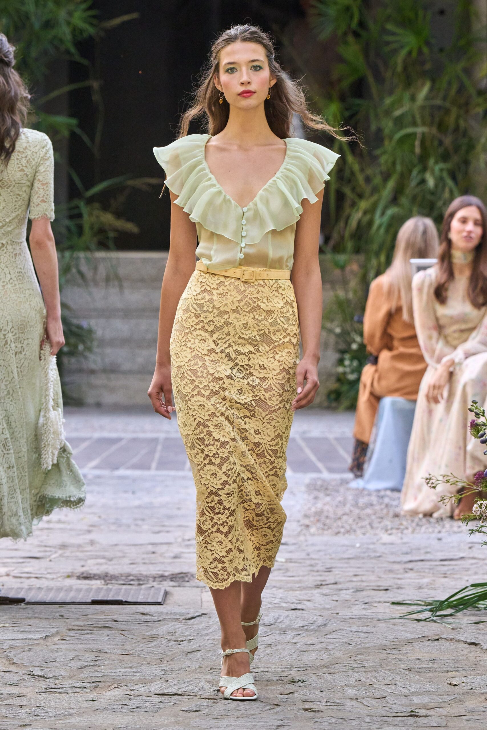 Pale pistachio chiffon blouse with a deep ruffled V and tiny buttons. Narrow matching belt with a small covered buckle. Buttercream floral lace pencil skirt, scalloped at the calf. Ivory ankle-strap sandals with crossed front bands. Small gold drop earrings. Garden wedding reception, the cake already softening in the heat.
