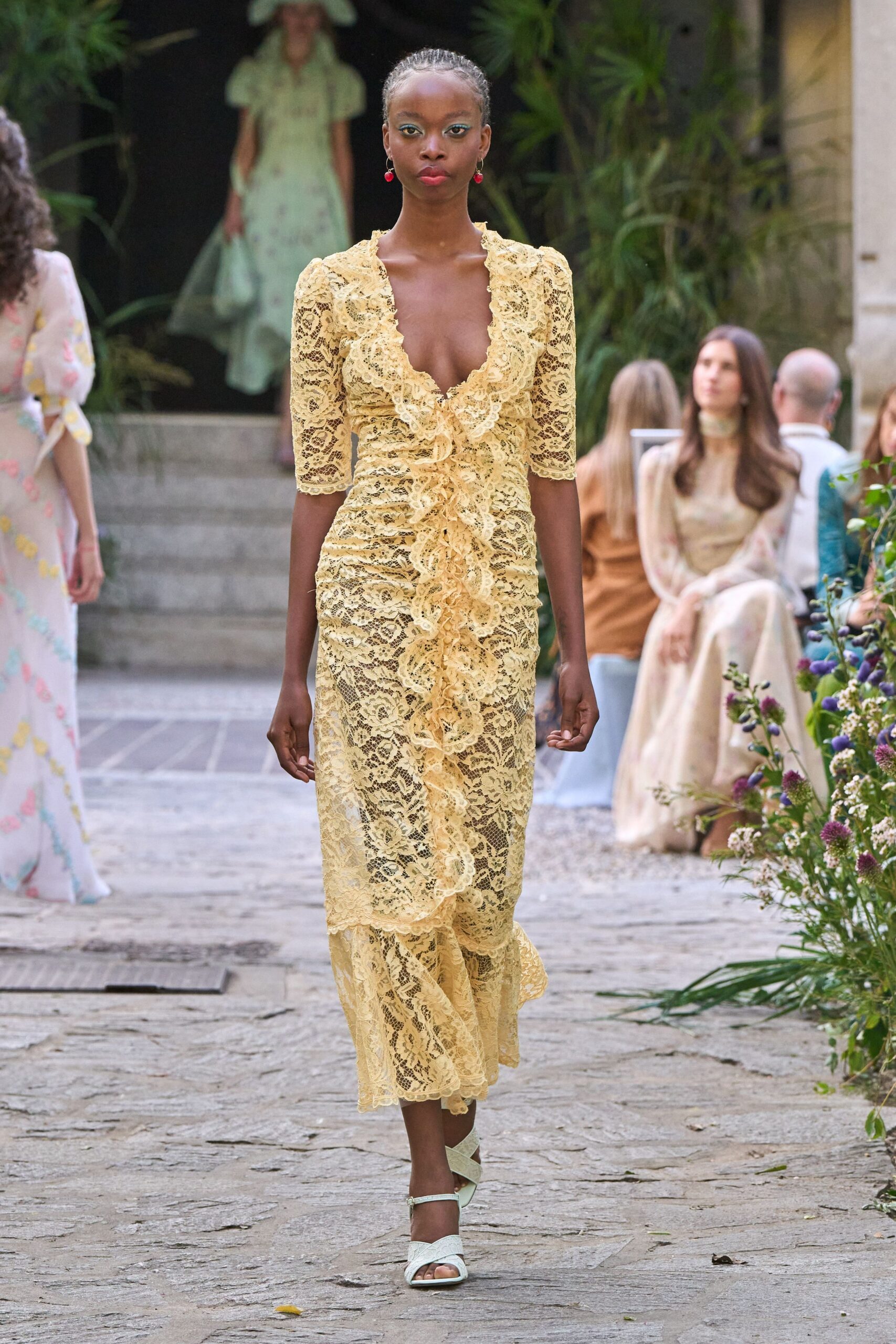 Buttercream floral lace dress, plunged neckline edged with layered ruffles and a rippling front flounce. Cherry-red drop earrings. White sandals with broad crisscross straps. Garden wedding at the hour the stone still gives back heat.