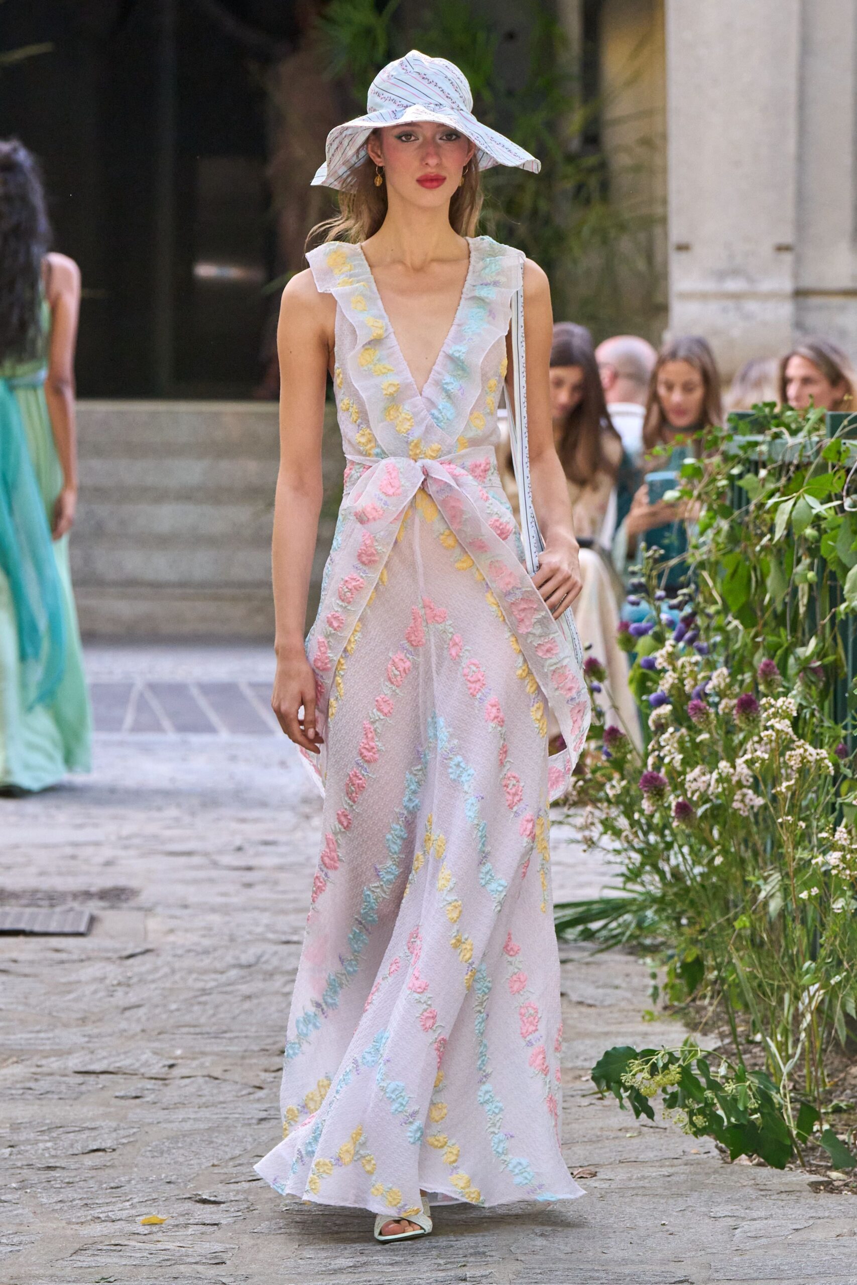 White cotton sunhat scribbled with red and blue ruled lines. Sheer lilac floor-length gown with ruffled plunge and pastel tufted sprigs. Wide self sash knotted low, tails falling over the skirt. Pale mint square-toe sandals. Garden wedding heat, crossing the gravel before the vows start.