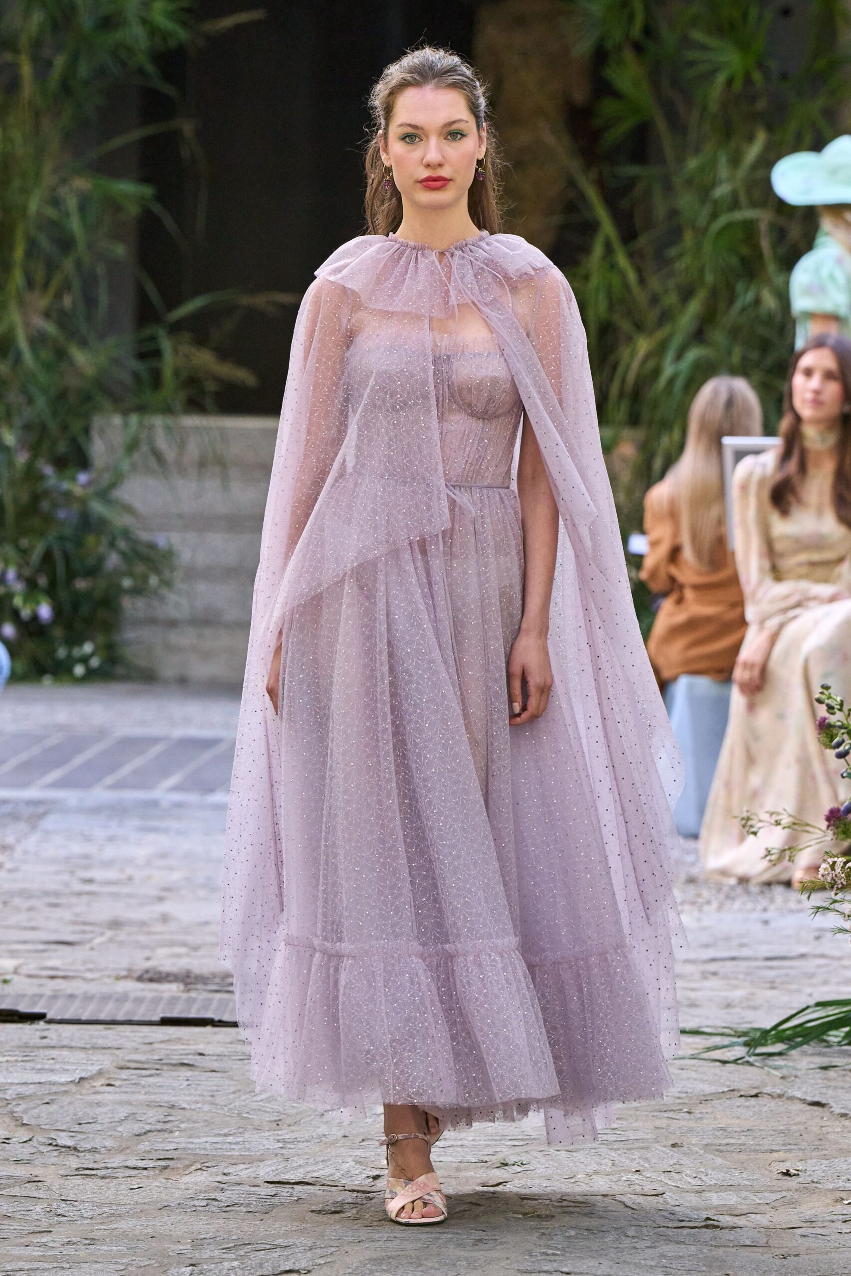 Sheer lilac tulle cape dusted with glitter, gathered into a ruffled neckline. Matching empire dress with corseted bodice and tiered ankle-length skirt. Blush satin sandals with slim ankle straps. Garden wedding at dusk, the air still warm off the stone path.