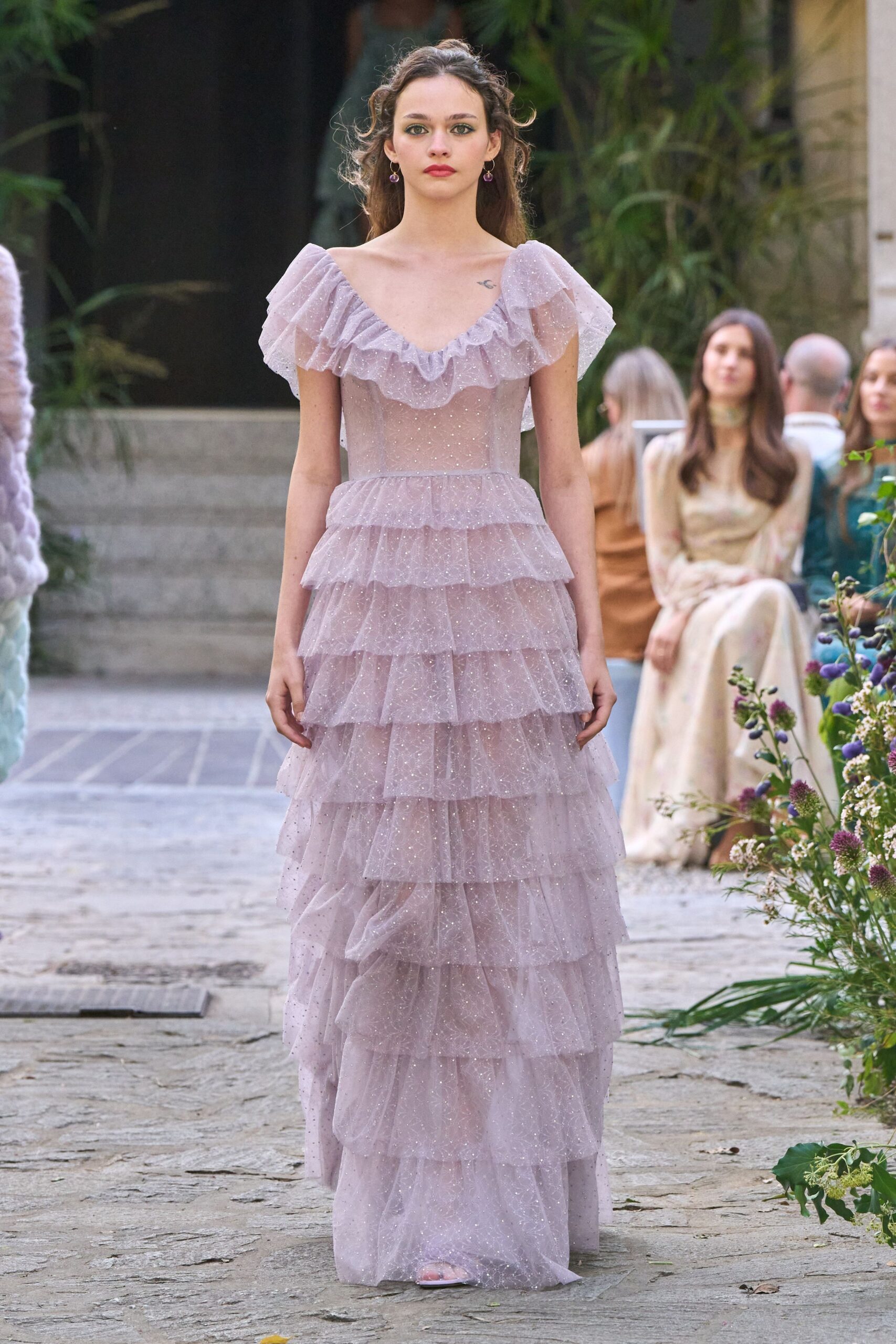 Lavender glitter tulle gown with a deep ruffled neckline and stacked tiered skirt. Small drop earrings with plum bead pendants. Pale open-toe sandals barely peeking beneath the hem. Garden wedding at dusk, before anyone asks you to make a speech.
