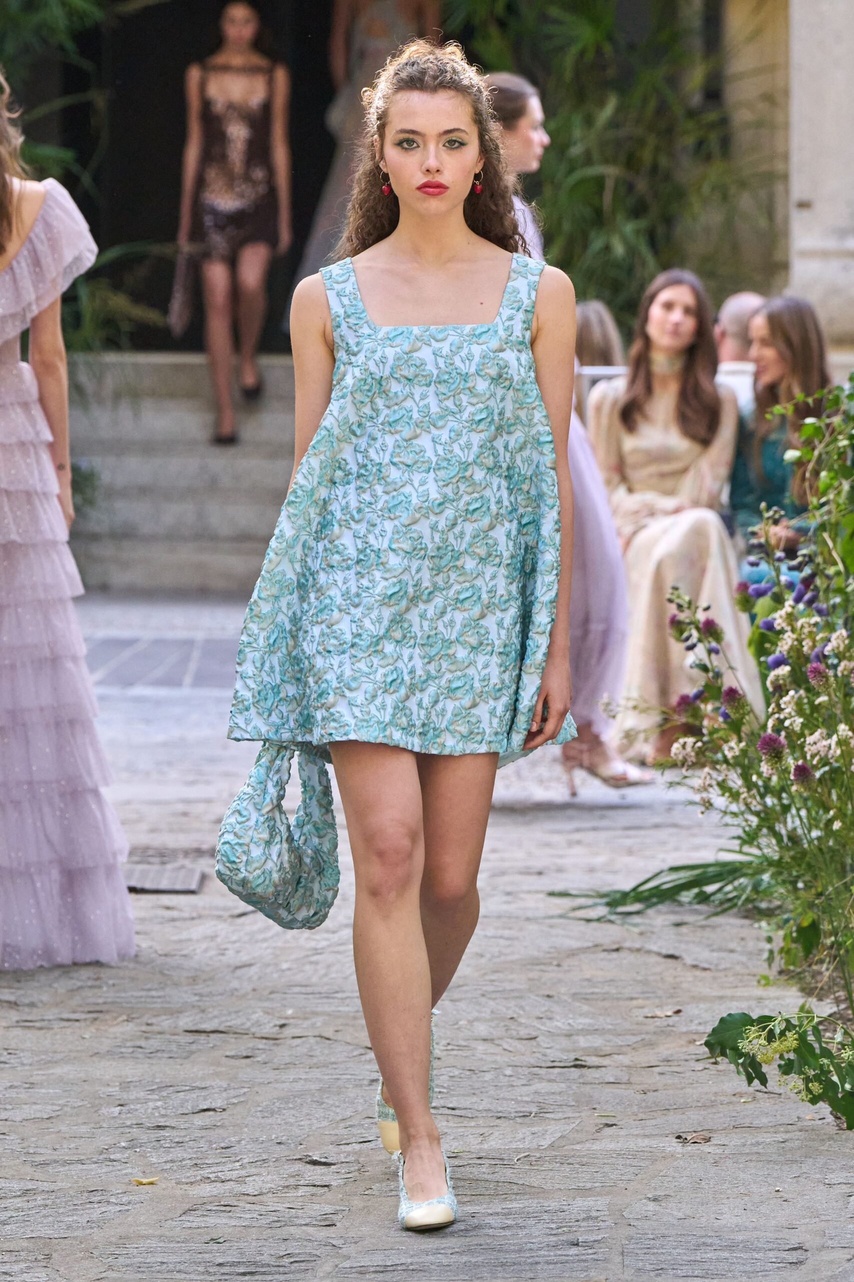 Pale aqua brocade mini dress, square neckline and puffed away from the body. Matching slouch bag in the same raised floral cloth. Cream pumps with aqua brocade vamps. Tiny cherry-drop earrings. Garden wedding spillover, champagne already warm on the stone.