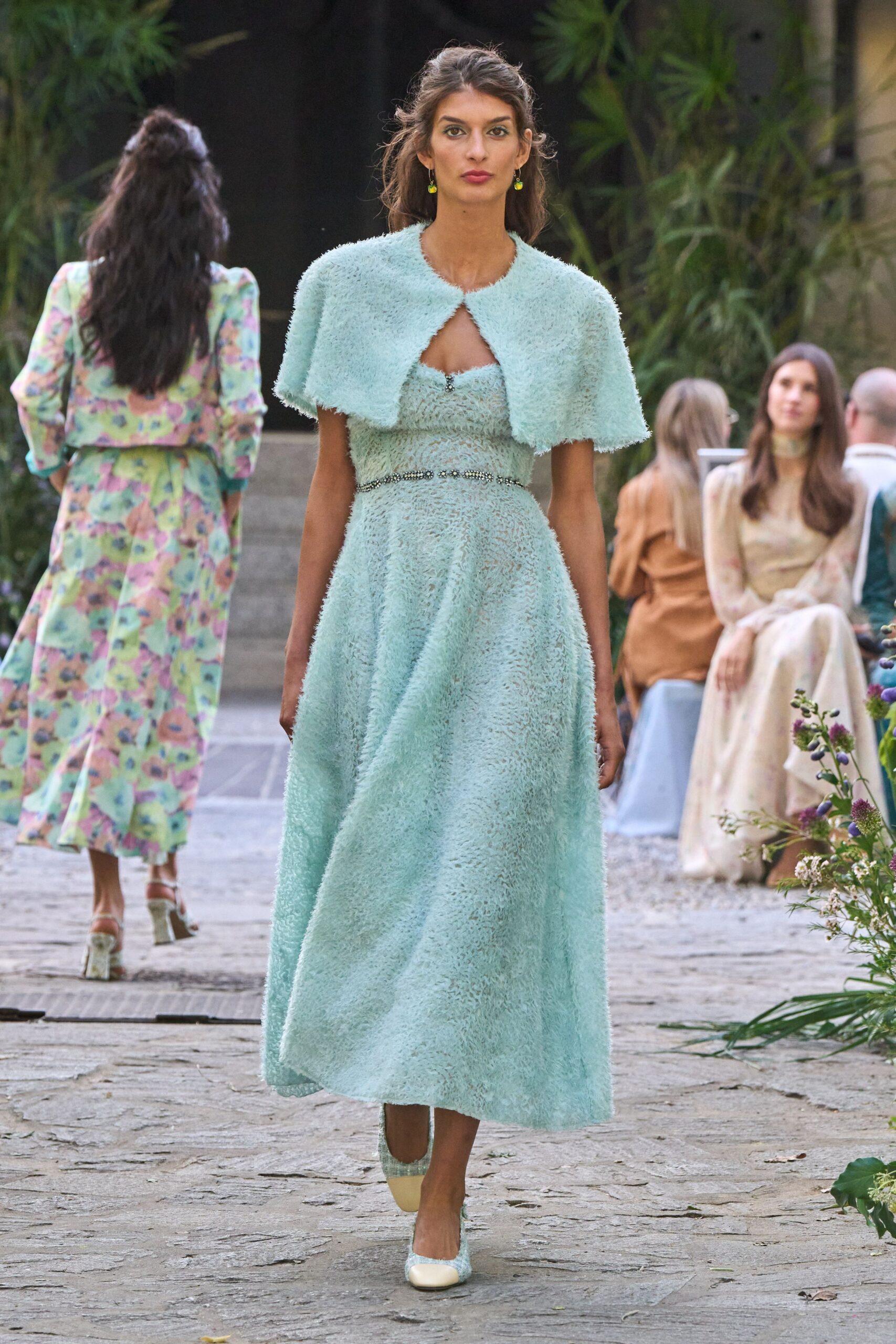 Powder-blue fuzzy knit capelet cut short with a single front clasp. Matching midi dress in airy eyelash texture, jeweled trim cinching the waist. Tiny lime drop earrings. Cream-toe pumps in the same pale fuzzy weave. Garden ceremony aisle, the heat still sitting in the stone.