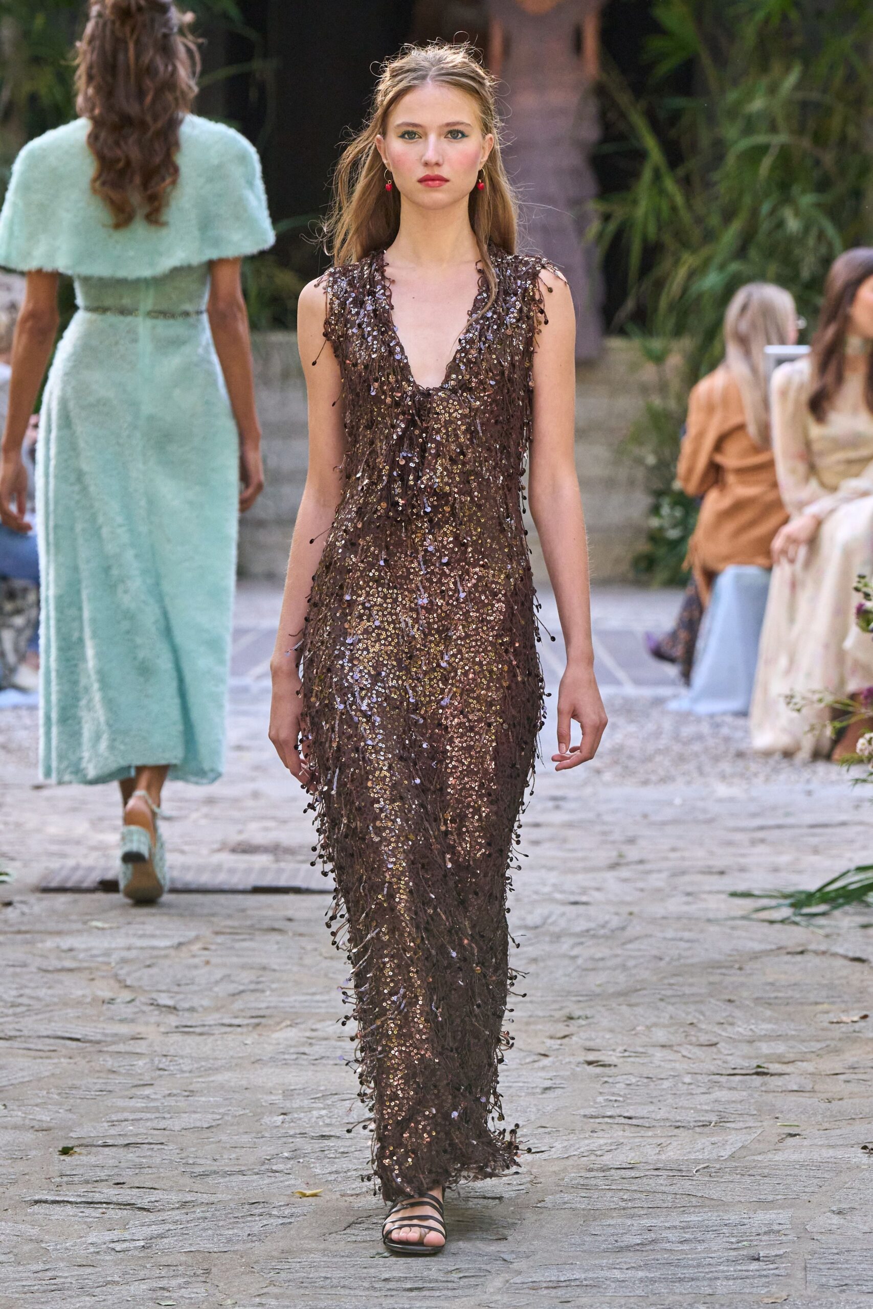 Small red drop earrings. Deep V column gown crusted in bronze sequins and beaded fringe. Black strappy flat sandals, barely visible under the hem. Crossing the garden path after dinner while the candles burn down.
