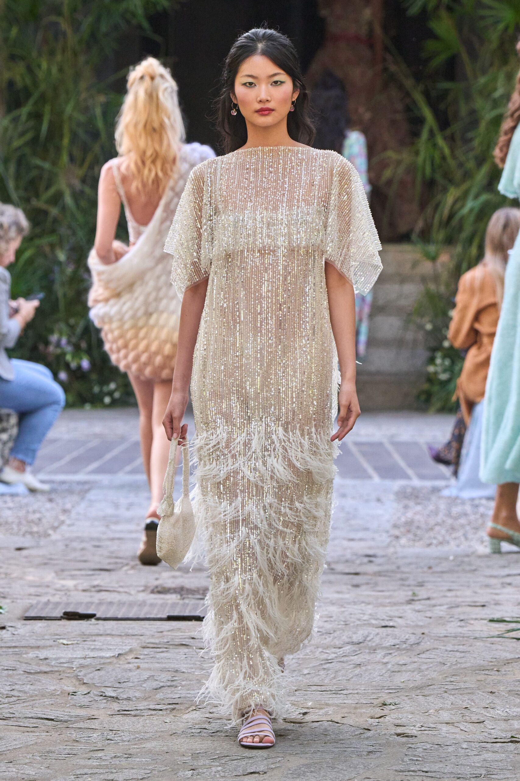 Ivory beaded caftan, vertical sequin strands over a sheer column. Ostrich fringe erupts at the hem and skims the calves. Small ivory beaded pouch with a narrow loop handle. Pale lilac straps crossing low over the toes. Garden stones still warm after sunset, heading back from a wedding by the sea.