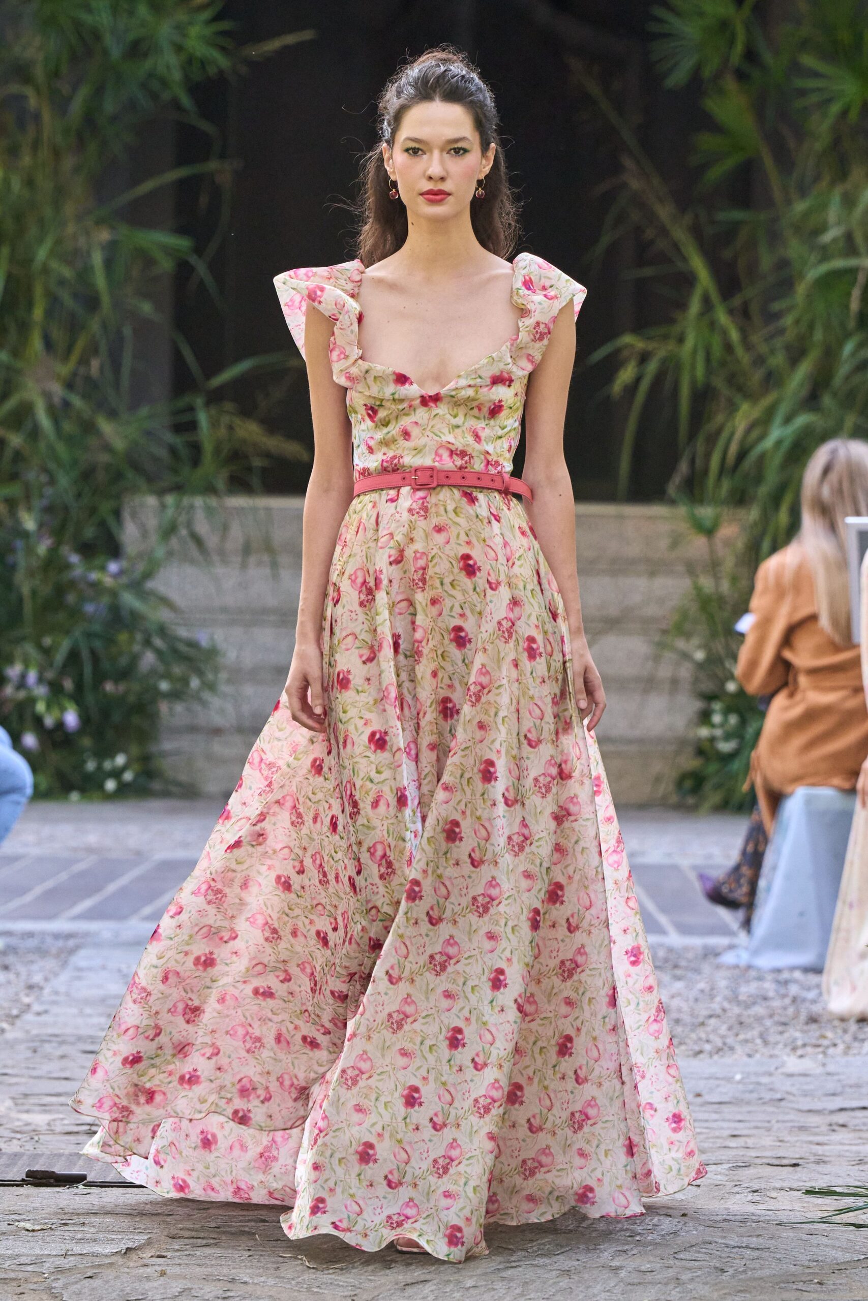 Cream floral gown with ruffled cap sleeves and a scalloped sweetheart neckline. Slim rose leather belt, the buckle set just off center. Small drop earrings with dark garnet beads. Garden wedding before sunset, gravel catching in the hem.