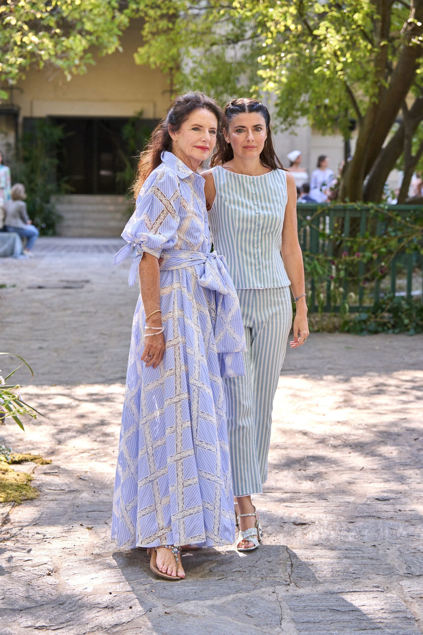 Powder-blue striped shell top with tiny scallops edging the armholes. Matching straight trousers in the same narrow vertical stripe. White ankle-strap sandals with a pale floral vamp. Walking through a shaded courtyard after the ceremony let out.
