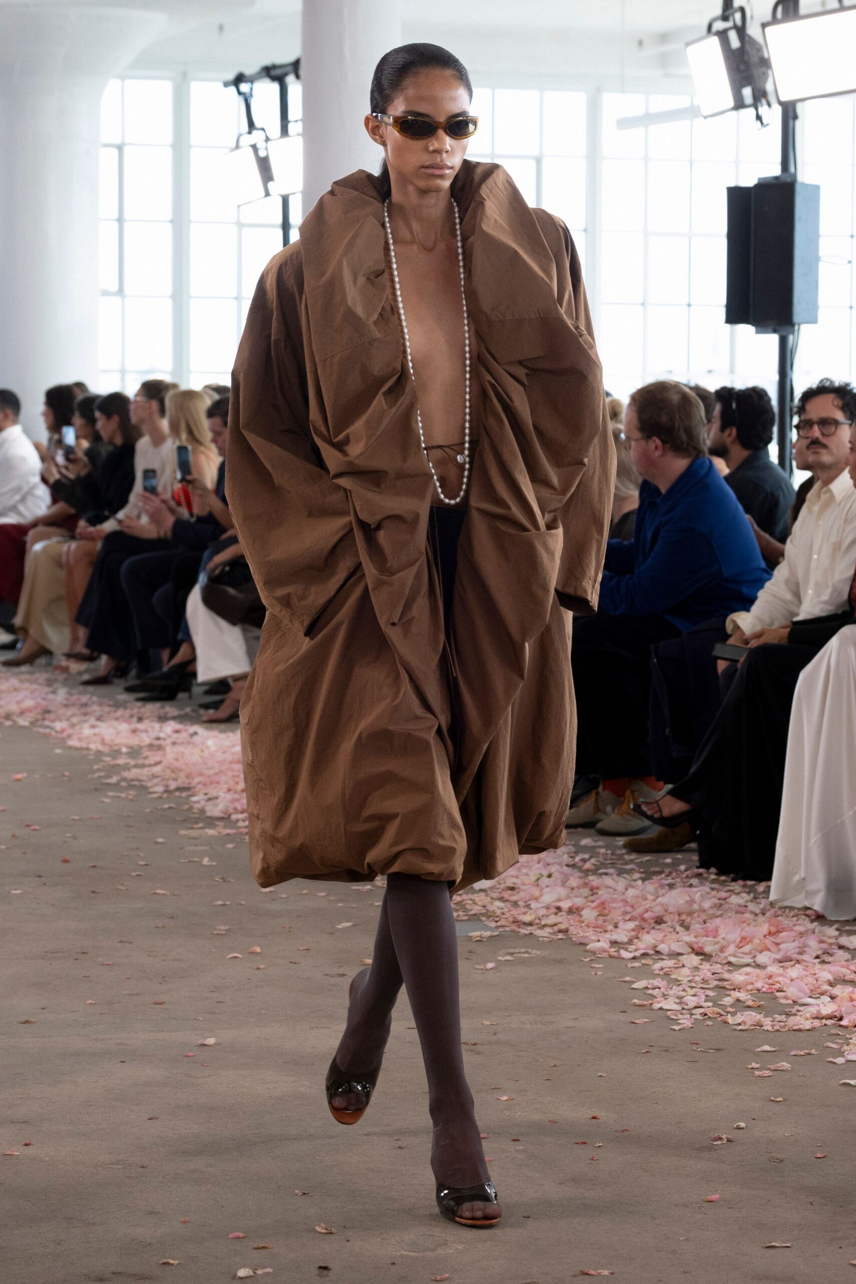 Narrow amber frames with dark oval lenses. Voluminous brown taffeta cocoon coat, crushed into huge standing folds. Long pearl strand dropping to the bare waist. Chocolate sheer tights. Black patent peep-toe heels with a raw wood sole edge. For crossing a hotel lobby at dawn with yesterday's flowers underfoot.