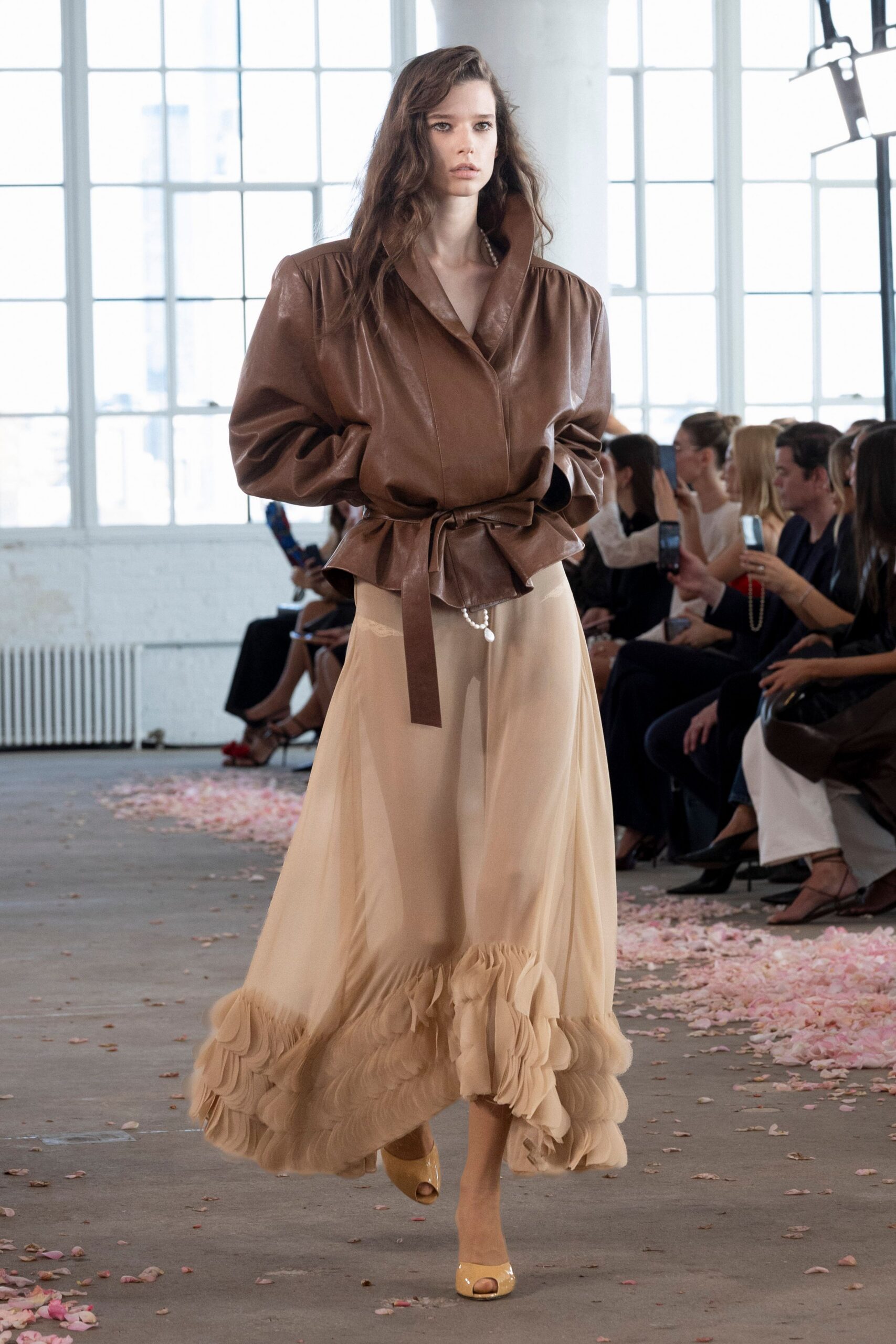 Cognac leather jacket with inflated shoulders and a self-tie peplum waist. Nude chiffon skirt, pearl drop peeking at the hip, hem built from ruffled tiers. Cream patent peep-toe pumps. Crossing a loft floor strewn with petals after the ceremony emptied out.