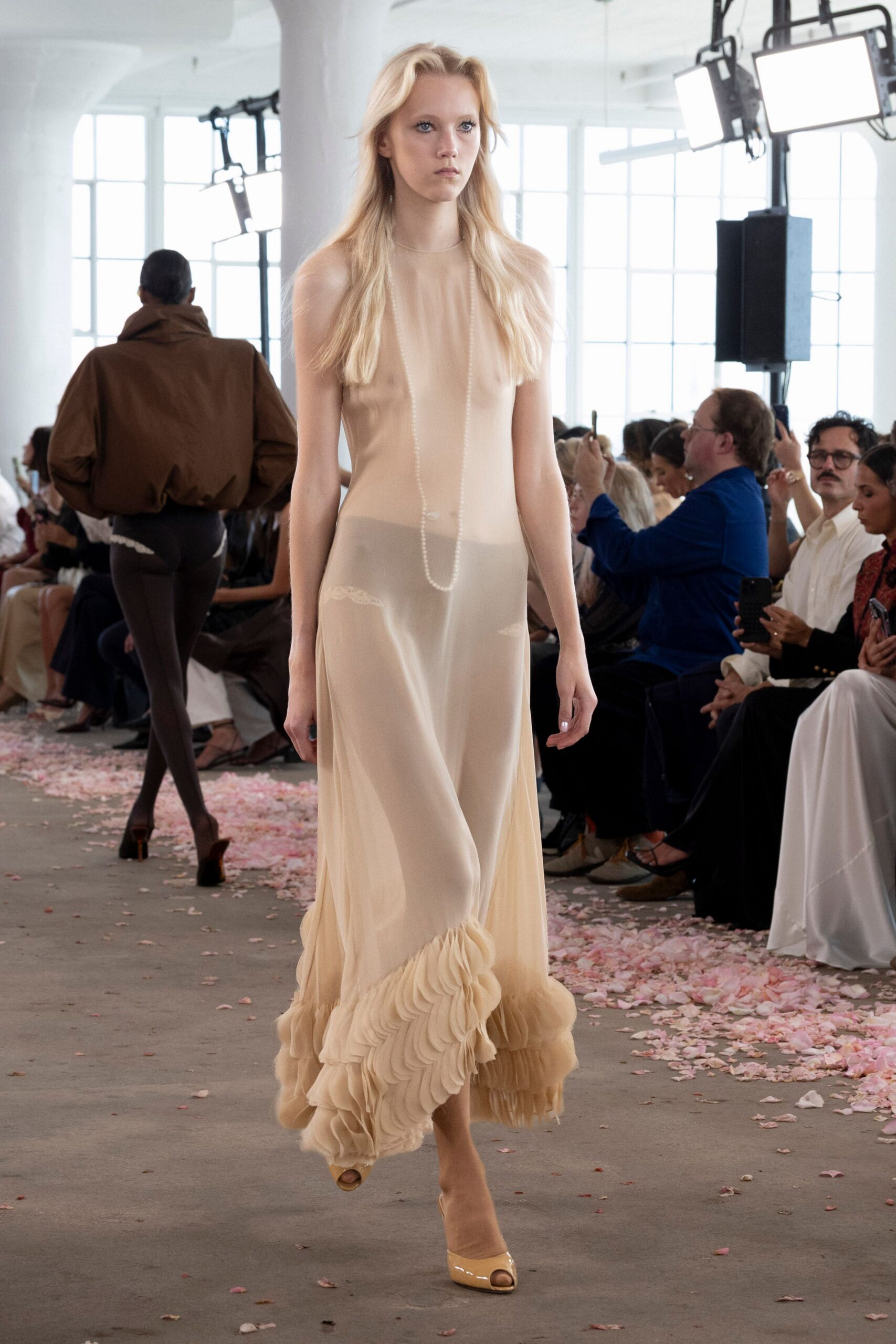 Nude sheer slip dress, long pearl strand falling over the torso. Greyed nude underlayer with small lace insets showing at the hips. Tiered chiffon flounces bunching thickly around the ankle. Cream patent peep-toes. After the ceremony, crossing a floor still sticky with crushed petals.